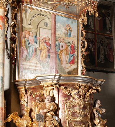 Zdjęcie nr 2: Ambona przyścienna, wisząca, z wejściem od tyłu dwubiegowymi schodami z podestem. Kosz na rzucie owalu o wklęsłych ściankach bocznych i wypukłej frontowej, na wypukłym cokole zdobionym od frontu płaskorzeźbionym motywem rocaille’u. U spodu kosza pięć kabłąków dekorowanych u góry palmetami i fragmentami rocaille’ów, zakończonych zwisem w formie winnego grona. Ścianki kosza opięte wklęsłym pilastrami; od frontu z figurami św. Jana i św. Mateusza siedzących na obłokach, trzymających otwarte księgi z napisem: OVI EX DEO EST VERBA DEI AUDIT / PROPTEREA VOS NON AUDITIS, QVIA EX DEO NON ESTIS. Joanne 8,47; oraz: ERAT ENIM / DOCENS EOS / SICUT PO=/TESTATEM /HABENS / ET NON / SICUT / SCRIBAE / EORUM / ET PHARI=/SAEI. Mat 7,18; na tle pozostałych pilastrów symbole ewangelistów: orzeł trzymający w ‘szponach kałamarz oraz głowa wołu. Ścianki w polach dekorowane rocaille’ami, frontowa z różanymi gałązkami. Kosz zwieńczony profilowanym gzymsem, występującym nad pilastrami. Na zaplecku obraz Szymona Kawalskiego Rozesłanie Apostołów, w kształcie leżącego prostokąta, w profilowanej, złoconej ramie, ujęty uszami utworzonymi z motywów rocaille’ów. Baldachim powtarza rzut kosza, wsparty na belkowaniu dekorowanym palmetami i rocaille’ami w partii fryzu, z podwieszonym lambrekinem. Powyżej ażurowa konstrukcja z wolut dekorowanych rocaille’ami, zbliżona do korony, otaczająca półpostać Chrystusa trzymającego jabłko królewskie i laskę pasterską. U spodu baldachimu podwieszona Gołębica Ducha Świętego w glorii. Struktura marmoryzowana laserunkowo, profile srebrzone, ornamenty złocone.