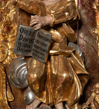 Zdjęcie nr 10: Ambona przyścienna, wisząca, z wejściem od tyłu dwubiegowymi schodami z podestem. Kosz na rzucie owalu o wklęsłych ściankach bocznych i wypukłej frontowej, na wypukłym cokole zdobionym od frontu płaskorzeźbionym motywem rocaille’u. U spodu kosza pięć kabłąków dekorowanych u góry palmetami i fragmentami rocaille’ów, zakończonych zwisem w formie winnego grona. Ścianki kosza opięte wklęsłym pilastrami; od frontu z figurami św. Jana i św. Mateusza siedzących na obłokach, trzymających otwarte księgi z napisem: OVI EX DEO EST VERBA DEI AUDIT / PROPTEREA VOS NON AUDITIS, QVIA EX DEO NON ESTIS. Joanne 8,47; oraz: ERAT ENIM / DOCENS EOS / SICUT PO=/TESTATEM /HABENS / ET NON / SICUT / SCRIBAE / EORUM / ET PHARI=/SAEI. Mat 7,18; na tle pozostałych pilastrów symbole ewangelistów: orzeł trzymający w ‘szponach kałamarz oraz głowa wołu. Ścianki w polach dekorowane rocaille’ami, frontowa z różanymi gałązkami. Kosz zwieńczony profilowanym gzymsem, występującym nad pilastrami. Na zaplecku obraz Szymona Kawalskiego Rozesłanie Apostołów, w kształcie leżącego prostokąta, w profilowanej, złoconej ramie, ujęty uszami utworzonymi z motywów rocaille’ów. Baldachim powtarza rzut kosza, wsparty na belkowaniu dekorowanym palmetami i rocaille’ami w partii fryzu, z podwieszonym lambrekinem. Powyżej ażurowa konstrukcja z wolut dekorowanych rocaille’ami, zbliżona do korony, otaczająca półpostać Chrystusa trzymającego jabłko królewskie i laskę pasterską. U spodu baldachimu podwieszona Gołębica Ducha Świętego w glorii. Struktura marmoryzowana laserunkowo, profile srebrzone, ornamenty złocone.