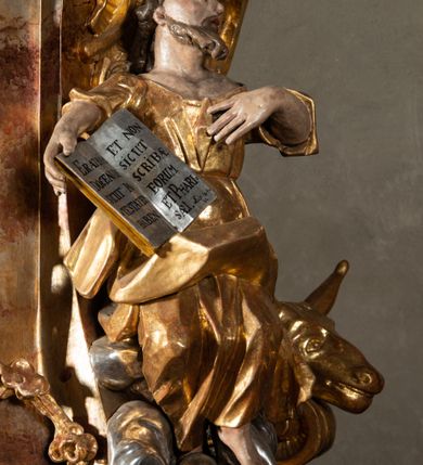 Zdjęcie nr 8: Ambona przyścienna, wisząca, z wejściem od tyłu dwubiegowymi schodami z podestem. Kosz na rzucie owalu o wklęsłych ściankach bocznych i wypukłej frontowej, na wypukłym cokole zdobionym od frontu płaskorzeźbionym motywem rocaille’u. U spodu kosza pięć kabłąków dekorowanych u góry palmetami i fragmentami rocaille’ów, zakończonych zwisem w formie winnego grona. Ścianki kosza opięte wklęsłym pilastrami; od frontu z figurami św. Jana i św. Mateusza siedzących na obłokach, trzymających otwarte księgi z napisem: OVI EX DEO EST VERBA DEI AUDIT / PROPTEREA VOS NON AUDITIS, QVIA EX DEO NON ESTIS. Joanne 8,47; oraz: ERAT ENIM / DOCENS EOS / SICUT PO=/TESTATEM /HABENS / ET NON / SICUT / SCRIBAE / EORUM / ET PHARI=/SAEI. Mat 7,18; na tle pozostałych pilastrów symbole ewangelistów: orzeł trzymający w ‘szponach kałamarz oraz głowa wołu. Ścianki w polach dekorowane rocaille’ami, frontowa z różanymi gałązkami. Kosz zwieńczony profilowanym gzymsem, występującym nad pilastrami. Na zaplecku obraz Szymona Kawalskiego Rozesłanie Apostołów, w kształcie leżącego prostokąta, w profilowanej, złoconej ramie, ujęty uszami utworzonymi z motywów rocaille’ów. Baldachim powtarza rzut kosza, wsparty na belkowaniu dekorowanym palmetami i rocaille’ami w partii fryzu, z podwieszonym lambrekinem. Powyżej ażurowa konstrukcja z wolut dekorowanych rocaille’ami, zbliżona do korony, otaczająca półpostać Chrystusa trzymającego jabłko królewskie i laskę pasterską. U spodu baldachimu podwieszona Gołębica Ducha Świętego w glorii. Struktura marmoryzowana laserunkowo, profile srebrzone, ornamenty złocone.