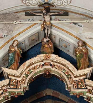 Zdjęcie nr 1: Belka profilowana, o formie pełnego belkowania, wygięta faliście, pośrodku wyłamana łukiem półkolistym nadwieszonym. Od frontu fryz dekorowany akantem i rozetami; od strony prezbiterium data: 1770; od spodu dekorowana rozetami. Na belce Grupa Ukrzyżowania. Rzeźby pełnoplastyczne, polichromowane. Pośrodku krzyż z figurą martwego Chrystusa. Ciało w lekkim zwisie, o szeroko rozłożonych ramionach. Kolana delikatnie ugięte, stopy przebite jednym gwoździem, założone prawa na lewą. Ciało wychudzone, smukłe, o podkreślonej muskulaturze, z zaznaczonymi śladami męki. Twarz pociągła, szczupła, oczy zamknięte; okolona zarostem. Włosy długie, opadające na ramiona. Na głowie korona cierniowa. Perizonium krótkie, ściśle opinające biodra, delikatnie drapowanie, ze zwisem tkaniny przy prawym boku. Na górnej belce krzyża titulus w formie kartusza z literami: INRI. U dołu krzyża klęcząca św. Maria Magdalena zwróconej en trois quarts w lewo, z głową skierowaną ku górze. Lewą ręką przytrzymuje stopę Chrystusa, prawa wyciągnięta przed siebie. Włosy upięte z tyłu głowy. Ubrana w bogatą suknię z gorsetem i dekoracyjnych rękawach oraz płaszcz zarzucony na ramiona. Po prawej stronie krzyża Matki Boska, ukazana frontalnie, z delikatnie pochylona głową i dłońmi złożonymi w geście modlitwy. Twarz pociągła, z uwypuklonym podbródkiem. Maria ubrana w suknię oraz płaszcz przewiązany na lewym ramieniu. Na głowie welon. Po lewej stronie krzyża św. Jan Ewangelista, ukazany en face, z głową skierowaną ku górze i dłońmi skrzyżowanymi na piersi. Twarz pociągła o młodzieńczych rysach, włosy długie, kręcone. Ubrany w długa tunikę przewiązaną w talii oraz płaszcz przewiązany na lewym ramieniu. Polichromia naturalistyczna w odsłoniętych partiach ciała, szaty złocone z laserunkiem.