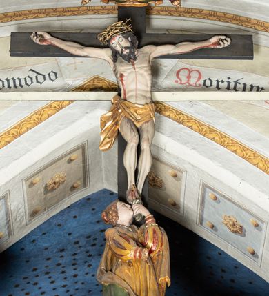 Zdjęcie nr 4: Belka profilowana, o formie pełnego belkowania, wygięta faliście, pośrodku wyłamana łukiem półkolistym nadwieszonym. Od frontu fryz dekorowany akantem i rozetami; od strony prezbiterium data: 1770; od spodu dekorowana rozetami. Na belce Grupa Ukrzyżowania. Rzeźby pełnoplastyczne, polichromowane. Pośrodku krzyż z figurą martwego Chrystusa. Ciało w lekkim zwisie, o szeroko rozłożonych ramionach. Kolana delikatnie ugięte, stopy przebite jednym gwoździem, założone prawa na lewą. Ciało wychudzone, smukłe, o podkreślonej muskulaturze, z zaznaczonymi śladami męki. Twarz pociągła, szczupła, oczy zamknięte; okolona zarostem. Włosy długie, opadające na ramiona. Na głowie korona cierniowa. Perizonium krótkie, ściśle opinające biodra, delikatnie drapowanie, ze zwisem tkaniny przy prawym boku. Na górnej belce krzyża titulus w formie kartusza z literami: INRI. U dołu krzyża klęcząca św. Maria Magdalena zwróconej en trois quarts w lewo, z głową skierowaną ku górze. Lewą ręką przytrzymuje stopę Chrystusa, prawa wyciągnięta przed siebie. Włosy upięte z tyłu głowy. Ubrana w bogatą suknię z gorsetem i dekoracyjnych rękawach oraz płaszcz zarzucony na ramiona. Po prawej stronie krzyża Matki Boska, ukazana frontalnie, z delikatnie pochylona głową i dłońmi złożonymi w geście modlitwy. Twarz pociągła, z uwypuklonym podbródkiem. Maria ubrana w suknię oraz płaszcz przewiązany na lewym ramieniu. Na głowie welon. Po lewej stronie krzyża św. Jan Ewangelista, ukazany en face, z głową skierowaną ku górze i dłońmi skrzyżowanymi na piersi. Twarz pociągła o młodzieńczych rysach, włosy długie, kręcone. Ubrany w długa tunikę przewiązaną w talii oraz płaszcz przewiązany na lewym ramieniu. Polichromia naturalistyczna w odsłoniętych partiach ciała, szaty złocone z laserunkiem.