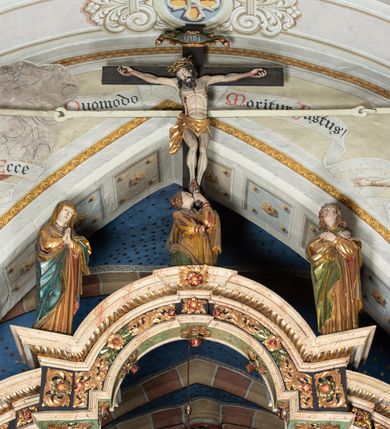 Zdjęcie nr 2: Belka profilowana, o formie pełnego belkowania, wygięta faliście, pośrodku wyłamana łukiem półkolistym nadwieszonym. Od frontu fryz dekorowany akantem i rozetami; od strony prezbiterium data: 1770; od spodu dekorowana rozetami. Na belce Grupa Ukrzyżowania. Rzeźby pełnoplastyczne, polichromowane. Pośrodku krzyż z figurą martwego Chrystusa. Ciało w lekkim zwisie, o szeroko rozłożonych ramionach. Kolana delikatnie ugięte, stopy przebite jednym gwoździem, założone prawa na lewą. Ciało wychudzone, smukłe, o podkreślonej muskulaturze, z zaznaczonymi śladami męki. Twarz pociągła, szczupła, oczy zamknięte; okolona zarostem. Włosy długie, opadające na ramiona. Na głowie korona cierniowa. Perizonium krótkie, ściśle opinające biodra, delikatnie drapowanie, ze zwisem tkaniny przy prawym boku. Na górnej belce krzyża titulus w formie kartusza z literami: INRI. U dołu krzyża klęcząca św. Maria Magdalena zwróconej en trois quarts w lewo, z głową skierowaną ku górze. Lewą ręką przytrzymuje stopę Chrystusa, prawa wyciągnięta przed siebie. Włosy upięte z tyłu głowy. Ubrana w bogatą suknię z gorsetem i dekoracyjnych rękawach oraz płaszcz zarzucony na ramiona. Po prawej stronie krzyża Matki Boska, ukazana frontalnie, z delikatnie pochylona głową i dłońmi złożonymi w geście modlitwy. Twarz pociągła, z uwypuklonym podbródkiem. Maria ubrana w suknię oraz płaszcz przewiązany na lewym ramieniu. Na głowie welon. Po lewej stronie krzyża św. Jan Ewangelista, ukazany en face, z głową skierowaną ku górze i dłońmi skrzyżowanymi na piersi. Twarz pociągła o młodzieńczych rysach, włosy długie, kręcone. Ubrany w długa tunikę przewiązaną w talii oraz płaszcz przewiązany na lewym ramieniu. Polichromia naturalistyczna w odsłoniętych partiach ciała, szaty złocone z laserunkiem.