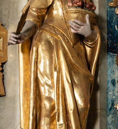 Zdjęcie nr 2: Rzeźba pełnoplastyczna, przyścienna, z tyłu drążona, ustawiona na niskim postumencie na rzucie kwadratu. Figura ukazana bokiem w stosunku do widza, zwrócona w lewo ku środkowi ołtarza, stojąca w kontrapoście, o esowato wygiętej sylwetce. W lewej, wyciągniętej w bok ręce trzyma palmę męczeństwa, prawą przytula do piersi kosz z różami. Twarz okrągła, szeroka, niewielkie oczy i nos, usta delikatnie rozchylone, wydatny podbródek. Włosy upięte z tyłu, na głowie korona otwarta. Ubrana w długą, delikatnie drapowaną, ściśle przylegająca do ciała suknię z krótkimi rękawami, gorset oraz płaszcz, delikatnie drapowany, przewiązany na piersi, opadający na plecy i upięty na lewym biodrze. Polichromia naturalistyczna w odsłoniętych partiach ciała, szaty i atrybuty złocone.