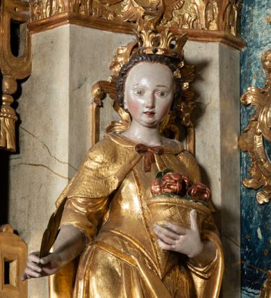 Zdjęcie nr 1: Rzeźba pełnoplastyczna, przyścienna, z tyłu drążona, ustawiona na niskim postumencie na rzucie kwadratu. Figura ukazana bokiem w stosunku do widza, zwrócona w lewo ku środkowi ołtarza, stojąca w kontrapoście, o esowato wygiętej sylwetce. W lewej, wyciągniętej w bok ręce trzyma palmę męczeństwa, prawą przytula do piersi kosz z różami. Twarz okrągła, szeroka, niewielkie oczy i nos, usta delikatnie rozchylone, wydatny podbródek. Włosy upięte z tyłu, na głowie korona otwarta. Ubrana w długą, delikatnie drapowaną, ściśle przylegająca do ciała suknię z krótkimi rękawami, gorset oraz płaszcz, delikatnie drapowany, przewiązany na piersi, opadający na plecy i upięty na lewym biodrze. Polichromia naturalistyczna w odsłoniętych partiach ciała, szaty i atrybuty złocone.