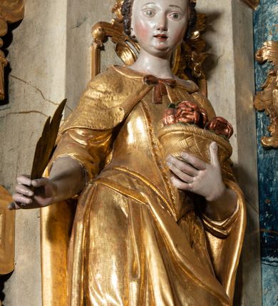Zdjęcie nr 3: Rzeźba pełnoplastyczna, przyścienna, z tyłu drążona, ustawiona na niskim postumencie na rzucie kwadratu. Figura ukazana bokiem w stosunku do widza, zwrócona w lewo ku środkowi ołtarza, stojąca w kontrapoście, o esowato wygiętej sylwetce. W lewej, wyciągniętej w bok ręce trzyma palmę męczeństwa, prawą przytula do piersi kosz z różami. Twarz okrągła, szeroka, niewielkie oczy i nos, usta delikatnie rozchylone, wydatny podbródek. Włosy upięte z tyłu, na głowie korona otwarta. Ubrana w długą, delikatnie drapowaną, ściśle przylegająca do ciała suknię z krótkimi rękawami, gorset oraz płaszcz, delikatnie drapowany, przewiązany na piersi, opadający na plecy i upięty na lewym biodrze. Polichromia naturalistyczna w odsłoniętych partiach ciała, szaty i atrybuty złocone.