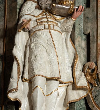 Zdjęcie nr 2: Rzeźba pełnoplastyczna, przyścienna, z tyłu drążona, na niskim postumencie.  Figura zwrócona en face, o delikatnie skręconej, przechylonej w tył sylwetce; głowa skierowana w lewą stronę, delikatnie uniesiona. Lewą ręką ugiętą, wyciągniętą przed siebie, podtrzymuje poduszkę z koroną zamkniętą; prawą ugiętą, wyciągniętą w bok, trzyma berło. Twarz okrągła, oczy zmrużone, patrzące do góry, nos długi, prosty, usta delikatnie rozchylone; okolona długą brodą o skręconych puklach, opadająca na pierś. Włosy długie, z przedziałkiem na środku głowy, skręcone w grube pukle. Święty ubrany w spodnie i wysokie buty; kaftan z wzorzystej, płaskorzeźbionej tkaniny, zapinany na guzy, z pasem i złoconą lamówką; płaszcz spięty na piersi, opadający przez ramiona na boki, o zawijających się brzegach. Polichromia naturalistyczna w odsłoniętych partiach ciała, szaty białe, atrybuty i detale stroju złocone.