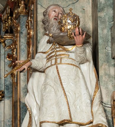 Zdjęcie nr 1: Rzeźba pełnoplastyczna, przyścienna, z tyłu drążona, na niskim postumencie.  Figura zwrócona en face, o delikatnie skręconej, przechylonej w tył sylwetce; głowa skierowana w lewą stronę, delikatnie uniesiona. Lewą ręką ugiętą, wyciągniętą przed siebie, podtrzymuje poduszkę z koroną zamkniętą; prawą ugiętą, wyciągniętą w bok, trzyma berło. Twarz okrągła, oczy zmrużone, patrzące do góry, nos długi, prosty, usta delikatnie rozchylone; okolona długą brodą o skręconych puklach, opadająca na pierś. Włosy długie, z przedziałkiem na środku głowy, skręcone w grube pukle. Święty ubrany w spodnie i wysokie buty; kaftan z wzorzystej, płaskorzeźbionej tkaniny, zapinany na guzy, z pasem i złoconą lamówką; płaszcz spięty na piersi, opadający przez ramiona na boki, o zawijających się brzegach. Polichromia naturalistyczna w odsłoniętych partiach ciała, szaty białe, atrybuty i detale stroju złocone.