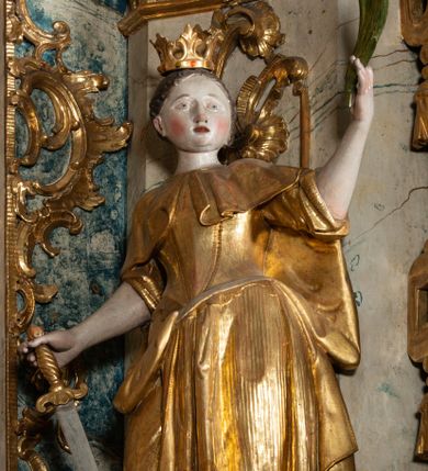 Zdjęcie nr 1: Rzeźba pełnoplastyczna, przyścienna, z tyłu drążona, ustawiona na niskim postumencie na rzucie kwadratu. Święta, ukazana w en trois quatrs, zwrócona w prawo, z głową skierowaną w lewo, stoi w kontrapoście, delikatnie przechylona do tyłu. W lewej, uniesionej ręce trzyma palmę męczeństwa, w prawej miecz, skierowany ostrzem w dół. Przy jej prawym boku koło. Twarz okrągła, szeroka, niewielkie oczy i nos, usta delikatnie rozchylone. Włosy upięte z tyłu, na głowie korona otwarta. Ubrana w długą suknię spodnią z krótkimi rękawami, suknie wierzchnią z gorsetem, sięgającą kolan dekorowaną rytymi pasami z koronkowym brzegiem, oraz płaszcz, delikatnie drapowany, przewiązany na piersi, opadający na plecy i upięty na prawym biodrze. Polichromia naturalistyczna w odsłoniętych partiach ciała, szaty i atrybuty złocone.