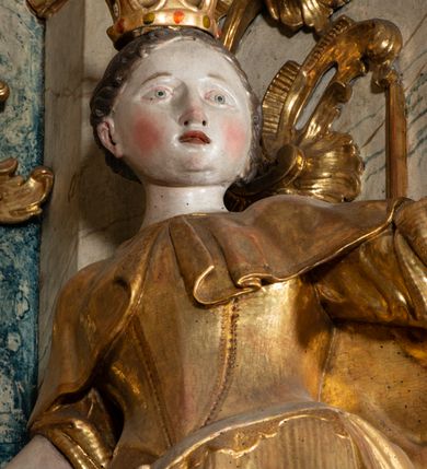 Zdjęcie nr 2: Rzeźba pełnoplastyczna, przyścienna, z tyłu drążona, ustawiona na niskim postumencie na rzucie kwadratu. Święta, ukazana w en trois quatrs, zwrócona w prawo, z głową skierowaną w lewo, stoi w kontrapoście, delikatnie przechylona do tyłu. W lewej, uniesionej ręce trzyma palmę męczeństwa, w prawej miecz, skierowany ostrzem w dół. Przy jej prawym boku koło. Twarz okrągła, szeroka, niewielkie oczy i nos, usta delikatnie rozchylone. Włosy upięte z tyłu, na głowie korona otwarta. Ubrana w długą suknię spodnią z krótkimi rękawami, suknie wierzchnią z gorsetem, sięgającą kolan dekorowaną rytymi pasami z koronkowym brzegiem, oraz płaszcz, delikatnie drapowany, przewiązany na piersi, opadający na plecy i upięty na prawym biodrze. Polichromia naturalistyczna w odsłoniętych partiach ciała, szaty i atrybuty złocone.