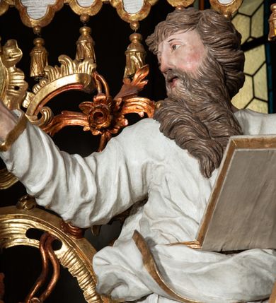 Zdjęcie nr 2: Rzeźba pełnoplastyczna, przyścienna, z tyłu drążona. Święty ukazany frontalnie, z głową zwróconą w prawo, w kierunku środka ołtarza. Prawa ręka uniesiona, wyciągnięta w bok, lewą, ugiętą, podtrzymuje opartą na biodrze zamkniętą księgę i miecz zwrócony ostrzem w dół. Twarz pociągła, o starczych rysach, oczy niewielkie, długi prosty nos, usta rozchylone; okolona długą brodą o skręconych puklach, opadającą na pierś. Włosy krótkie, gęste, kręcone. Święty obrany w tunikę obficie marszczoną w partii torsu oraz płaszcz, gęsto drapowany w grube fałdy, przewieszony przez lewe ramię i opadający na kolana. Polichromia naturalistyczna w odsłoniętych partiach ciała, szaty białe, atrybuty i detale stroju złocone.