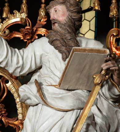 Zdjęcie nr 4: Rzeźba pełnoplastyczna, przyścienna, z tyłu drążona. Święty ukazany frontalnie, z głową zwróconą w prawo, w kierunku środka ołtarza. Prawa ręka uniesiona, wyciągnięta w bok, lewą, ugiętą, podtrzymuje opartą na biodrze zamkniętą księgę i miecz zwrócony ostrzem w dół. Twarz pociągła, o starczych rysach, oczy niewielkie, długi prosty nos, usta rozchylone; okolona długą brodą o skręconych puklach, opadającą na pierś. Włosy krótkie, gęste, kręcone. Święty obrany w tunikę obficie marszczoną w partii torsu oraz płaszcz, gęsto drapowany w grube fałdy, przewieszony przez lewe ramię i opadający na kolana. Polichromia naturalistyczna w odsłoniętych partiach ciała, szaty białe, atrybuty i detale stroju złocone.