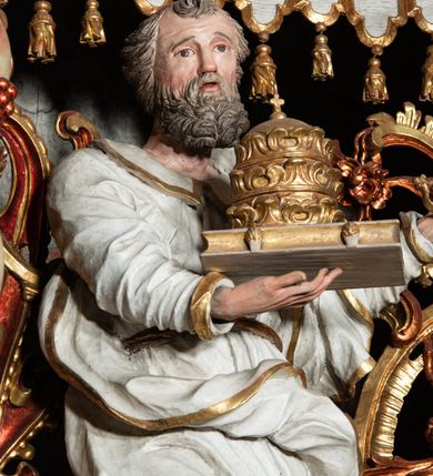 Zdjęcie nr 2: Rzeźba pełnoplastyczna, przyścienna, z tyłu, drążona, ustawiona na rzeźbionym fragmencie skały. Święty ukazany frontalnie, z prawą nogą ugiętą, opartą o skałę. Prawą rękę podtrzymuje przed sobą zamkniętą księgę z potrójną papieską tiarą; w lewej, wyciągniętej w bok ręce trzyma dwa skrzyżowane klucze. Twarz pociągła, o starczych rysach, niewielkich oczach, prostym długim nosie, niewielkich, rozchylonych ustach, okolona krótką, kręconą brodą. Włosy krótkie, kręcone, pukle po bokach i na czubku głowy. Święty ubrany w tunikę z kołnierzem, przepasaną w talii, marszczoną w gęste drobne fałdki, oraz płaszcz przerzucony przez lewe ramię i opadający gęstymi, grubymi fałdami, skręconej i ekspresyjnie rozwianej tkaniny na kolana i prawy bok. Polichromia naturalistyczna w odsłoniętych partiach ciała, szaty białe, atrybuty i detale stroju złocone.