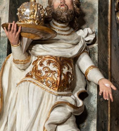 Zdjęcie nr 2: Rzeźba pełnoplastyczna, przyścienna, z tyłu drążona, ustawiona na niskiej prostopadłościennej podstawie. Święty, ukazany frontalnie, stoi w kontrapoście, z głową odchyloną do tyłu i wzrokiem skierowanym ku górze.  W prawej, uniesionej ręce trzyma tacę z zamkniętą koroną; lewa wyciągnięta przed siebie. Twarz pociągła, oczy małe, zwrócone w górę, nos prosty, usta rozchylone; okolona krótką, kręconą brodą. Włosy półdługie, układające się w grube pukle, z przedziałkiem na środku głowy. Święty ubrany w spodnie i wysokie buty; tunikę z długim rękawem, delikatnie drapowaną, sięgającą kolan, ze złotą lamówką; puklerz dekorowany płaskorzeźbionym akantem; płaszcz obficie drapowany, o zawiniętych brzegach, zawiązany na lewym ramieniu, opadający na plecy i zawinięty na lewej nodze. Polichromia naturalistyczna w odsłoniętych partiach ciała, szaty białe, atrybuty i detale stroju złocone.