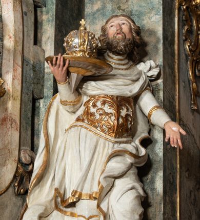 Zdjęcie nr 1: Rzeźba pełnoplastyczna, przyścienna, z tyłu drążona, ustawiona na niskiej prostopadłościennej podstawie. Święty, ukazany frontalnie, stoi w kontrapoście, z głową odchyloną do tyłu i wzrokiem skierowanym ku górze.  W prawej, uniesionej ręce trzyma tacę z zamkniętą koroną; lewa wyciągnięta przed siebie. Twarz pociągła, oczy małe, zwrócone w górę, nos prosty, usta rozchylone; okolona krótką, kręconą brodą. Włosy półdługie, układające się w grube pukle, z przedziałkiem na środku głowy. Święty ubrany w spodnie i wysokie buty; tunikę z długim rękawem, delikatnie drapowaną, sięgającą kolan, ze złotą lamówką; puklerz dekorowany płaskorzeźbionym akantem; płaszcz obficie drapowany, o zawiniętych brzegach, zawiązany na lewym ramieniu, opadający na plecy i zawinięty na lewej nodze. Polichromia naturalistyczna w odsłoniętych partiach ciała, szaty białe, atrybuty i detale stroju złocone.