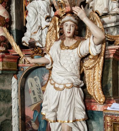 Zdjęcie nr 1: Rzeźby pełnoplastyczne, przyścienne, z tyłu drążone. Figury analogiczne, zwrócone antytetycznie. Anioły ukazane en face, stojące w kontrapoście, o lekko esowato wygiętych sylwetkach, na głowach podtrzymują woluty, ręce od wewnętrznej strony ołtarza, ugięte, uniesione do góry, podtrzymują woluty, zewnętrzne wyciągnięte w bok, dzierżą miecze o płomienistych ostrzach. Twarze pociągłe, o niewielkich oczach i długich prostych nosach. Włosy długie, opadające gęstymi puklami na ramiona. Anioły ubrane w białe tuniki sięgające do kolan, ze złotą lamówką, szatę spodnią sięgającą do kostek, odsłaniającą prawe nogi, obficie drapowane w grube fałdy, ekspresyjnie poruszone; łuskowy puklerz oraz przewieszony ukośnie przez prawe ramię płaszcz. Na głowach hełm, na nogach wysokie sandały. Skrzydła złożone.Polichromia naturalistyczna w odsłoniętych partiach ciała, szaty białe, detale i skrzydła złocone.