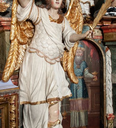 Zdjęcie nr 2: Rzeźby pełnoplastyczne, przyścienne, z tyłu drążone. Figury analogiczne, zwrócone antytetycznie. Anioły ukazane en face, stojące w kontrapoście, o lekko esowato wygiętych sylwetkach, na głowach podtrzymują woluty, ręce od wewnętrznej strony ołtarza, ugięte, uniesione do góry, podtrzymują woluty, zewnętrzne wyciągnięte w bok, dzierżą miecze o płomienistych ostrzach. Twarze pociągłe, o niewielkich oczach i długich prostych nosach. Włosy długie, opadające gęstymi puklami na ramiona. Anioły ubrane w białe tuniki sięgające do kolan, ze złotą lamówką, szatę spodnią sięgającą do kostek, odsłaniającą prawe nogi, obficie drapowane w grube fałdy, ekspresyjnie poruszone; łuskowy puklerz oraz przewieszony ukośnie przez prawe ramię płaszcz. Na głowach hełm, na nogach wysokie sandały. Skrzydła złożone.Polichromia naturalistyczna w odsłoniętych partiach ciała, szaty białe, detale i skrzydła złocone.