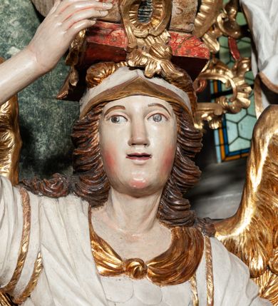 Zdjęcie nr 8: Rzeźby pełnoplastyczne, przyścienne, z tyłu drążone. Figury analogiczne, zwrócone antytetycznie. Anioły ukazane en face, stojące w kontrapoście, o lekko esowato wygiętych sylwetkach, na głowach podtrzymują woluty, ręce od wewnętrznej strony ołtarza, ugięte, uniesione do góry, podtrzymują woluty, zewnętrzne wyciągnięte w bok, dzierżą miecze o płomienistych ostrzach. Twarze pociągłe, o niewielkich oczach i długich prostych nosach. Włosy długie, opadające gęstymi puklami na ramiona. Anioły ubrane w białe tuniki sięgające do kolan, ze złotą lamówką, szatę spodnią sięgającą do kostek, odsłaniającą prawe nogi, obficie drapowane w grube fałdy, ekspresyjnie poruszone; łuskowy puklerz oraz przewieszony ukośnie przez prawe ramię płaszcz. Na głowach hełm, na nogach wysokie sandały. Skrzydła złożone.Polichromia naturalistyczna w odsłoniętych partiach ciała, szaty białe, detale i skrzydła złocone.