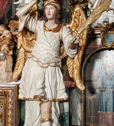 Zdjęcie nr 6: Rzeźby pełnoplastyczne, przyścienne, z tyłu drążone. Figury analogiczne, zwrócone antytetycznie. Anioły ukazane en face, stojące w kontrapoście, o lekko esowato wygiętych sylwetkach, na głowach podtrzymują woluty, ręce od wewnętrznej strony ołtarza, ugięte, uniesione do góry, podtrzymują woluty, zewnętrzne wyciągnięte w bok, dzierżą miecze o płomienistych ostrzach. Twarze pociągłe, o niewielkich oczach i długich prostych nosach. Włosy długie, opadające gęstymi puklami na ramiona. Anioły ubrane w białe tuniki sięgające do kolan, ze złotą lamówką, szatę spodnią sięgającą do kostek, odsłaniającą prawe nogi, obficie drapowane w grube fałdy, ekspresyjnie poruszone; łuskowy puklerz oraz przewieszony ukośnie przez prawe ramię płaszcz. Na głowach hełm, na nogach wysokie sandały. Skrzydła złożone.Polichromia naturalistyczna w odsłoniętych partiach ciała, szaty białe, detale i skrzydła złocone.