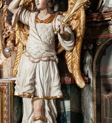 Zdjęcie nr 5: Rzeźby pełnoplastyczne, przyścienne, z tyłu drążone. Figury analogiczne, zwrócone antytetycznie. Anioły ukazane en face, stojące w kontrapoście, o lekko esowato wygiętych sylwetkach, na głowach podtrzymują woluty, ręce od wewnętrznej strony ołtarza, ugięte, uniesione do góry, podtrzymują woluty, zewnętrzne wyciągnięte w bok, dzierżą miecze o płomienistych ostrzach. Twarze pociągłe, o niewielkich oczach i długich prostych nosach. Włosy długie, opadające gęstymi puklami na ramiona. Anioły ubrane w białe tuniki sięgające do kolan, ze złotą lamówką, szatę spodnią sięgającą do kostek, odsłaniającą prawe nogi, obficie drapowane w grube fałdy, ekspresyjnie poruszone; łuskowy puklerz oraz przewieszony ukośnie przez prawe ramię płaszcz. Na głowach hełm, na nogach wysokie sandały. Skrzydła złożone.Polichromia naturalistyczna w odsłoniętych partiach ciała, szaty białe, detale i skrzydła złocone.