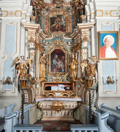 Zdjęcie nr 2: Ołtarz architektoniczny, jednoosiowy, jednokondygnacyjny ze zwieńczeniem, na cokole. Mensa drewniana umieszczona na drewnianej tumbie o wklęsło-wypukłych bokach. Antependium dekorowane rocaille’owym kartuszem z rytym gorejącym sercem, brzegi zdobione rocaille’em. W predelli przeszklona trumienka z figurą św. Jana Nepomucena, w profilowanej, złoconej ramie, na rocaillowej podstawie. Nastawa ujęta parami występujących do przodu filarów: wewnętrzne filary opięte pilastrami; zewnętrzne filary z wolutami zamiast trzonów, z figurami aniołów. Podpory dźwigają pełne belkowanie, przerwane nad polem środkowym. W polu nisza w kształcie stojącego prostokąta zamkniętego trójbocznie, w której obraz Święta Rodzina w profilowanej ramie. Przy filarach od wewnątrz rocaille’owe konsole z figurami św. Doroty i św. Katarzyny Aleksandryjskiej. Zwieńczenie w formie aedikuli zbliżonej do kwadratu o fantazyjnym wklęsło-wypukłym zamknięciu; ujęte pilastrami przechodzącymi w spływy wolutowe, opadające na belkowanie, tworzące postumenty dla figur św. Stefana i św. Emeryka. W polu zwieńczenia obraz z przedstawieniem Świętej Rodziny przy pracy, w kształcie kwadratu o wklęsłych narożach, w profilowanej ramie. Zwieńczenie zamknięte glorią promienistą z literami IHS w otoczeniu obłoków; na osi pilastrów rokokowe wazony z różami.W partii cokołów kanony w rocaille’owych ramach. Trzony pilastrów i woluty dekorowane rocaille’owymi płycinami, we fryzie rocaille. Nisza w polu środkowym ujęta rocaille’ami, zwieńczona mitrą książęcą, flankowana przez parę uskrzydlonych główek anielskich. Woluty zwieńczenia dekorowane motywami rocaille’ów. Rama obrazu w polu ujęta małżowiną, ornamentem roślinnym i rocaille’ami. Struktura marmoryzowana w odcieniu jasnoniebieskim, podpory, tumba i cokoły w odcieniu kości słoniowej; ornamentyka i profile złocone.