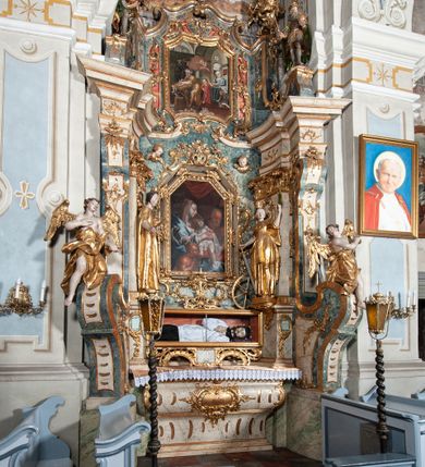 Zdjęcie nr 4: Ołtarz architektoniczny, jednoosiowy, jednokondygnacyjny ze zwieńczeniem, na cokole. Mensa drewniana umieszczona na drewnianej tumbie o wklęsło-wypukłych bokach. Antependium dekorowane rocaille’owym kartuszem z rytym gorejącym sercem, brzegi zdobione rocaille’em. W predelli przeszklona trumienka z figurą św. Jana Nepomucena, w profilowanej, złoconej ramie, na rocaillowej podstawie. Nastawa ujęta parami występujących do przodu filarów: wewnętrzne filary opięte pilastrami; zewnętrzne filary z wolutami zamiast trzonów, z figurami aniołów. Podpory dźwigają pełne belkowanie, przerwane nad polem środkowym. W polu nisza w kształcie stojącego prostokąta zamkniętego trójbocznie, w której obraz Święta Rodzina w profilowanej ramie. Przy filarach od wewnątrz rocaille’owe konsole z figurami św. Doroty i św. Katarzyny Aleksandryjskiej. Zwieńczenie w formie aedikuli zbliżonej do kwadratu o fantazyjnym wklęsło-wypukłym zamknięciu; ujęte pilastrami przechodzącymi w spływy wolutowe, opadające na belkowanie, tworzące postumenty dla figur św. Stefana i św. Emeryka. W polu zwieńczenia obraz z przedstawieniem Świętej Rodziny przy pracy, w kształcie kwadratu o wklęsłych narożach, w profilowanej ramie. Zwieńczenie zamknięte glorią promienistą z literami IHS w otoczeniu obłoków; na osi pilastrów rokokowe wazony z różami.W partii cokołów kanony w rocaille’owych ramach. Trzony pilastrów i woluty dekorowane rocaille’owymi płycinami, we fryzie rocaille. Nisza w polu środkowym ujęta rocaille’ami, zwieńczona mitrą książęcą, flankowana przez parę uskrzydlonych główek anielskich. Woluty zwieńczenia dekorowane motywami rocaille’ów. Rama obrazu w polu ujęta małżowiną, ornamentem roślinnym i rocaille’ami. Struktura marmoryzowana w odcieniu jasnoniebieskim, podpory, tumba i cokoły w odcieniu kości słoniowej; ornamentyka i profile złocone.