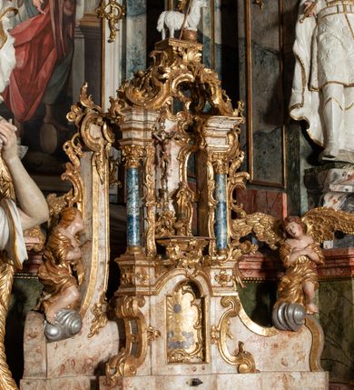 Zdjęcie nr 13: Ołtarz architektoniczny, trójosiowy, z wydzieloną częścią cokołową z bramkami bocznymi, jednokondygnacyjny, ze zwieńczeniem. Mensa drewniana z portatylem z marmuru dębnickiego, umieszczona na drewnianej, prostopadłościennej skrzyni.  Antepedium w kształcie leżącego prostokąta, ujęte profilowaną ramą dekorowaną liśćmi akantu i perełkowaniem. W polu płaskorzeźbione przedstawienie baranka leżącego na krzyżu, na obłoku, ujęte parą liści akantu oraz wicią roślinną z kwiatami i kiściami winogron. 
Na mensie tabernakulum z Tronem Łaski w formie prostopadłościanu na cokole, o wypukłej ściance frontowej, ujęte spływami wolutowymi podtrzymującymi niepełne belkowanie, wyłamane po środku łukiem półkolistym. Na froncie obrotowa nisza ujęta obramieniem w kształcie stojącego prostokąta zamkniętego łukiem nadwieszonym. W niszy ryte przedstawienie kielicha z Eucharystią w glorii, na obłoku, całość w rocaillowej ramie. Powyżej tron wystawienia z cokołem dekorowanym rocaille’em i parą ornamentalnych świeczników; ujęty parą kolumn dźwigających imposty. Podpory dżwigają rocaillowe kabłąki tworzące baldachim, na którego szczycie figurka baranka eucharystycznego leżącego na księdze. Ścianka w formie zbliżonej do stojącego prostokąta ujęta rocaille. Tabernakulum i tron ujęte okazałymi spływami wolutowymi z rocaillowymi uszami oraz na zakończeniach u dołu dwiema rzeźbami klęczących, adorujących aniołów.
Osie nastawy wydzielone w osiach skrajnych parą kolumn o skręconych trzonach i korynckich kapitelach umieszczonych na tle pilastrów, w osiach wewnętrznych parą pilastrów o trzonach w formie wolut z puttami z puttami, podtrzymywaną przez kariatydy w formie okazałych figur uskrzydlonych aniołów. Podpory wspierają pełne belkowanie z profilowanym gzymsem, występujące nad nimi, przerwane nad polem środkowym. W polu środkowym nisza ze sklepieniem kolebkowym wspartym na dwóch filarach i pilastrach. W głębi niszy obraz Św. Bartłomiej w profilowanej ramie w kształcie stojącego prostokąta zamkniętego łukiem odcinkowym. Na podniebiu niszy obraz Męczeństwo św. Bartłomieja w kształcie leżącego prostokąta ujętego niesymetrycznym obramieniem z motywem rocaille’ów. Nisza flankowana z figurami św. Stefana i św. Emeryka umieszczonymi na konsolach, na tle pilastrów. Pola retabulum w osiach skrajnych wklęsłe, między podporami prześwity z figurami św. Piotra i św. Pawła. W partii wysokiego cokołu bramki ujęte od zewnątrz wklęsłymi filarami podtrzymującymi belkowanie, wejścia z drzwiczkami w kształcie stojącego prostokąta zamkniętego łukiem półkolistym z obrazami przedstawiającymi Mojżesza i Aarona. Zwieńczenie w formie aedikuli ujętej pilastrami wspierającymi przerwane belkowanie oraz fantazyjnymi spływami wolutowymi, spływającymi na wewnętrzne podpory, przyjmując formę przerwanego przyczółka, na którym dwa putta i dwie figury siedzących aniołów. W osiach zewnętrznych podpór dwa putta siedzące kanelowanych postumentach i obłokach, prawe przytrzymuje skórę (atrybut św. Bartłomieja). W polu zwieńczenia obraz Św. Anna Samotrzeć w kształcie wydłużonego ośmioboku, w ornamentalnej ramie. Całość zwieńczona rzeźbioną półpostacią Boga Ojca pośród obłoków ukazanego w glorii promienistej. Drzwi do bramek flankowane liściastymi festonami, powyżej tarcze ujęte liśćmi laurowymi, zwieńczone koroną z klejnotem; lewa z klejnotem w postaci łabędzia z napisem: SOLI/ DEO/GLORIA, prawa z postacią ludzką z napisem: SOLI DEO HONOR. Woluty i fryz dekorowane motywami rocaille’ów oraz uskrzydlonymi główkami anielskimi. Osie boczne wypełnione ażurowymi rocaille’ami z gałązkami różanymi oraz ornamentem małżowinowo-chrząstkowym, belkowanie z podwieszonym lambrekinem z chwostami. Skręcone trzony kolumn zdobione wicią z winnej latorośli. Archiwolta nad polem środkowym dekorowana rocaille’em, rocaille’owym kartuszem i lambrekinem z chwostami. Woluty zwieńczenia, pilastry i naroża pola obrazowego dekorowane motywami rocaille’ów. 
Nastawa marmoryzowana w odcieniu jasnoszarym, profile marmoryzowane w kolorze jasnoróżowym, ornamenty i obramienia złocone.