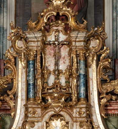 Zdjęcie nr 15: Ołtarz architektoniczny, trójosiowy, z wydzieloną częścią cokołową z bramkami bocznymi, jednokondygnacyjny, ze zwieńczeniem. Mensa drewniana z portatylem z marmuru dębnickiego, umieszczona na drewnianej, prostopadłościennej skrzyni.  Antepedium w kształcie leżącego prostokąta, ujęte profilowaną ramą dekorowaną liśćmi akantu i perełkowaniem. W polu płaskorzeźbione przedstawienie baranka leżącego na krzyżu, na obłoku, ujęte parą liści akantu oraz wicią roślinną z kwiatami i kiściami winogron. 
Na mensie tabernakulum z Tronem Łaski w formie prostopadłościanu na cokole, o wypukłej ściance frontowej, ujęte spływami wolutowymi podtrzymującymi niepełne belkowanie, wyłamane po środku łukiem półkolistym. Na froncie obrotowa nisza ujęta obramieniem w kształcie stojącego prostokąta zamkniętego łukiem nadwieszonym. W niszy ryte przedstawienie kielicha z Eucharystią w glorii, na obłoku, całość w rocaillowej ramie. Powyżej tron wystawienia z cokołem dekorowanym rocaille’em i parą ornamentalnych świeczników; ujęty parą kolumn dźwigających imposty. Podpory dżwigają rocaillowe kabłąki tworzące baldachim, na którego szczycie figurka baranka eucharystycznego leżącego na księdze. Ścianka w formie zbliżonej do stojącego prostokąta ujęta rocaille. Tabernakulum i tron ujęte okazałymi spływami wolutowymi z rocaillowymi uszami oraz na zakończeniach u dołu dwiema rzeźbami klęczących, adorujących aniołów.
Osie nastawy wydzielone w osiach skrajnych parą kolumn o skręconych trzonach i korynckich kapitelach umieszczonych na tle pilastrów, w osiach wewnętrznych parą pilastrów o trzonach w formie wolut z puttami z puttami, podtrzymywaną przez kariatydy w formie okazałych figur uskrzydlonych aniołów. Podpory wspierają pełne belkowanie z profilowanym gzymsem, występujące nad nimi, przerwane nad polem środkowym. W polu środkowym nisza ze sklepieniem kolebkowym wspartym na dwóch filarach i pilastrach. W głębi niszy obraz Św. Bartłomiej w profilowanej ramie w kształcie stojącego prostokąta zamkniętego łukiem odcinkowym. Na podniebiu niszy obraz Męczeństwo św. Bartłomieja w kształcie leżącego prostokąta ujętego niesymetrycznym obramieniem z motywem rocaille’ów. Nisza flankowana z figurami św. Stefana i św. Emeryka umieszczonymi na konsolach, na tle pilastrów. Pola retabulum w osiach skrajnych wklęsłe, między podporami prześwity z figurami św. Piotra i św. Pawła. W partii wysokiego cokołu bramki ujęte od zewnątrz wklęsłymi filarami podtrzymującymi belkowanie, wejścia z drzwiczkami w kształcie stojącego prostokąta zamkniętego łukiem półkolistym z obrazami przedstawiającymi Mojżesza i Aarona. Zwieńczenie w formie aedikuli ujętej pilastrami wspierającymi przerwane belkowanie oraz fantazyjnymi spływami wolutowymi, spływającymi na wewnętrzne podpory, przyjmując formę przerwanego przyczółka, na którym dwa putta i dwie figury siedzących aniołów. W osiach zewnętrznych podpór dwa putta siedzące kanelowanych postumentach i obłokach, prawe przytrzymuje skórę (atrybut św. Bartłomieja). W polu zwieńczenia obraz Św. Anna Samotrzeć w kształcie wydłużonego ośmioboku, w ornamentalnej ramie. Całość zwieńczona rzeźbioną półpostacią Boga Ojca pośród obłoków ukazanego w glorii promienistej. Drzwi do bramek flankowane liściastymi festonami, powyżej tarcze ujęte liśćmi laurowymi, zwieńczone koroną z klejnotem; lewa z klejnotem w postaci łabędzia z napisem: SOLI/ DEO/GLORIA, prawa z postacią ludzką z napisem: SOLI DEO HONOR. Woluty i fryz dekorowane motywami rocaille’ów oraz uskrzydlonymi główkami anielskimi. Osie boczne wypełnione ażurowymi rocaille’ami z gałązkami różanymi oraz ornamentem małżowinowo-chrząstkowym, belkowanie z podwieszonym lambrekinem z chwostami. Skręcone trzony kolumn zdobione wicią z winnej latorośli. Archiwolta nad polem środkowym dekorowana rocaille’em, rocaille’owym kartuszem i lambrekinem z chwostami. Woluty zwieńczenia, pilastry i naroża pola obrazowego dekorowane motywami rocaille’ów. 
Nastawa marmoryzowana w odcieniu jasnoszarym, profile marmoryzowane w kolorze jasnoróżowym, ornamenty i obramienia złocone.