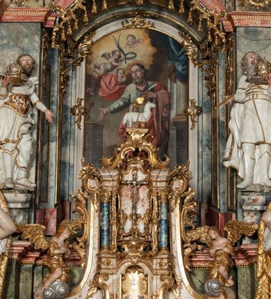 Zdjęcie nr 18: Ołtarz architektoniczny, trójosiowy, z wydzieloną częścią cokołową z bramkami bocznymi, jednokondygnacyjny, ze zwieńczeniem. Mensa drewniana z portatylem z marmuru dębnickiego, umieszczona na drewnianej, prostopadłościennej skrzyni.  Antepedium w kształcie leżącego prostokąta, ujęte profilowaną ramą dekorowaną liśćmi akantu i perełkowaniem. W polu płaskorzeźbione przedstawienie baranka leżącego na krzyżu, na obłoku, ujęte parą liści akantu oraz wicią roślinną z kwiatami i kiściami winogron. 
Na mensie tabernakulum z Tronem Łaski w formie prostopadłościanu na cokole, o wypukłej ściance frontowej, ujęte spływami wolutowymi podtrzymującymi niepełne belkowanie, wyłamane po środku łukiem półkolistym. Na froncie obrotowa nisza ujęta obramieniem w kształcie stojącego prostokąta zamkniętego łukiem nadwieszonym. W niszy ryte przedstawienie kielicha z Eucharystią w glorii, na obłoku, całość w rocaillowej ramie. Powyżej tron wystawienia z cokołem dekorowanym rocaille’em i parą ornamentalnych świeczników; ujęty parą kolumn dźwigających imposty. Podpory dżwigają rocaillowe kabłąki tworzące baldachim, na którego szczycie figurka baranka eucharystycznego leżącego na księdze. Ścianka w formie zbliżonej do stojącego prostokąta ujęta rocaille. Tabernakulum i tron ujęte okazałymi spływami wolutowymi z rocaillowymi uszami oraz na zakończeniach u dołu dwiema rzeźbami klęczących, adorujących aniołów.
Osie nastawy wydzielone w osiach skrajnych parą kolumn o skręconych trzonach i korynckich kapitelach umieszczonych na tle pilastrów, w osiach wewnętrznych parą pilastrów o trzonach w formie wolut z puttami z puttami, podtrzymywaną przez kariatydy w formie okazałych figur uskrzydlonych aniołów. Podpory wspierają pełne belkowanie z profilowanym gzymsem, występujące nad nimi, przerwane nad polem środkowym. W polu środkowym nisza ze sklepieniem kolebkowym wspartym na dwóch filarach i pilastrach. W głębi niszy obraz Św. Bartłomiej w profilowanej ramie w kształcie stojącego prostokąta zamkniętego łukiem odcinkowym. Na podniebiu niszy obraz Męczeństwo św. Bartłomieja w kształcie leżącego prostokąta ujętego niesymetrycznym obramieniem z motywem rocaille’ów. Nisza flankowana z figurami św. Stefana i św. Emeryka umieszczonymi na konsolach, na tle pilastrów. Pola retabulum w osiach skrajnych wklęsłe, między podporami prześwity z figurami św. Piotra i św. Pawła. W partii wysokiego cokołu bramki ujęte od zewnątrz wklęsłymi filarami podtrzymującymi belkowanie, wejścia z drzwiczkami w kształcie stojącego prostokąta zamkniętego łukiem półkolistym z obrazami przedstawiającymi Mojżesza i Aarona. Zwieńczenie w formie aedikuli ujętej pilastrami wspierającymi przerwane belkowanie oraz fantazyjnymi spływami wolutowymi, spływającymi na wewnętrzne podpory, przyjmując formę przerwanego przyczółka, na którym dwa putta i dwie figury siedzących aniołów. W osiach zewnętrznych podpór dwa putta siedzące kanelowanych postumentach i obłokach, prawe przytrzymuje skórę (atrybut św. Bartłomieja). W polu zwieńczenia obraz Św. Anna Samotrzeć w kształcie wydłużonego ośmioboku, w ornamentalnej ramie. Całość zwieńczona rzeźbioną półpostacią Boga Ojca pośród obłoków ukazanego w glorii promienistej. Drzwi do bramek flankowane liściastymi festonami, powyżej tarcze ujęte liśćmi laurowymi, zwieńczone koroną z klejnotem; lewa z klejnotem w postaci łabędzia z napisem: SOLI/ DEO/GLORIA, prawa z postacią ludzką z napisem: SOLI DEO HONOR. Woluty i fryz dekorowane motywami rocaille’ów oraz uskrzydlonymi główkami anielskimi. Osie boczne wypełnione ażurowymi rocaille’ami z gałązkami różanymi oraz ornamentem małżowinowo-chrząstkowym, belkowanie z podwieszonym lambrekinem z chwostami. Skręcone trzony kolumn zdobione wicią z winnej latorośli. Archiwolta nad polem środkowym dekorowana rocaille’em, rocaille’owym kartuszem i lambrekinem z chwostami. Woluty zwieńczenia, pilastry i naroża pola obrazowego dekorowane motywami rocaille’ów. 
Nastawa marmoryzowana w odcieniu jasnoszarym, profile marmoryzowane w kolorze jasnoróżowym, ornamenty i obramienia złocone.