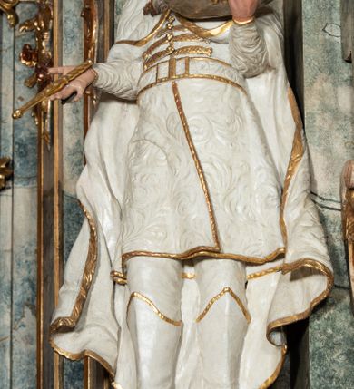 Zdjęcie nr 23: Ołtarz architektoniczny, trójosiowy, z wydzieloną częścią cokołową z bramkami bocznymi, jednokondygnacyjny, ze zwieńczeniem. Mensa drewniana z portatylem z marmuru dębnickiego, umieszczona na drewnianej, prostopadłościennej skrzyni.  Antepedium w kształcie leżącego prostokąta, ujęte profilowaną ramą dekorowaną liśćmi akantu i perełkowaniem. W polu płaskorzeźbione przedstawienie baranka leżącego na krzyżu, na obłoku, ujęte parą liści akantu oraz wicią roślinną z kwiatami i kiściami winogron. 
Na mensie tabernakulum z Tronem Łaski w formie prostopadłościanu na cokole, o wypukłej ściance frontowej, ujęte spływami wolutowymi podtrzymującymi niepełne belkowanie, wyłamane po środku łukiem półkolistym. Na froncie obrotowa nisza ujęta obramieniem w kształcie stojącego prostokąta zamkniętego łukiem nadwieszonym. W niszy ryte przedstawienie kielicha z Eucharystią w glorii, na obłoku, całość w rocaillowej ramie. Powyżej tron wystawienia z cokołem dekorowanym rocaille’em i parą ornamentalnych świeczników; ujęty parą kolumn dźwigających imposty. Podpory dżwigają rocaillowe kabłąki tworzące baldachim, na którego szczycie figurka baranka eucharystycznego leżącego na księdze. Ścianka w formie zbliżonej do stojącego prostokąta ujęta rocaille. Tabernakulum i tron ujęte okazałymi spływami wolutowymi z rocaillowymi uszami oraz na zakończeniach u dołu dwiema rzeźbami klęczących, adorujących aniołów.
Osie nastawy wydzielone w osiach skrajnych parą kolumn o skręconych trzonach i korynckich kapitelach umieszczonych na tle pilastrów, w osiach wewnętrznych parą pilastrów o trzonach w formie wolut z puttami z puttami, podtrzymywaną przez kariatydy w formie okazałych figur uskrzydlonych aniołów. Podpory wspierają pełne belkowanie z profilowanym gzymsem, występujące nad nimi, przerwane nad polem środkowym. W polu środkowym nisza ze sklepieniem kolebkowym wspartym na dwóch filarach i pilastrach. W głębi niszy obraz Św. Bartłomiej w profilowanej ramie w kształcie stojącego prostokąta zamkniętego łukiem odcinkowym. Na podniebiu niszy obraz Męczeństwo św. Bartłomieja w kształcie leżącego prostokąta ujętego niesymetrycznym obramieniem z motywem rocaille’ów. Nisza flankowana z figurami św. Stefana i św. Emeryka umieszczonymi na konsolach, na tle pilastrów. Pola retabulum w osiach skrajnych wklęsłe, między podporami prześwity z figurami św. Piotra i św. Pawła. W partii wysokiego cokołu bramki ujęte od zewnątrz wklęsłymi filarami podtrzymującymi belkowanie, wejścia z drzwiczkami w kształcie stojącego prostokąta zamkniętego łukiem półkolistym z obrazami przedstawiającymi Mojżesza i Aarona. Zwieńczenie w formie aedikuli ujętej pilastrami wspierającymi przerwane belkowanie oraz fantazyjnymi spływami wolutowymi, spływającymi na wewnętrzne podpory, przyjmując formę przerwanego przyczółka, na którym dwa putta i dwie figury siedzących aniołów. W osiach zewnętrznych podpór dwa putta siedzące kanelowanych postumentach i obłokach, prawe przytrzymuje skórę (atrybut św. Bartłomieja). W polu zwieńczenia obraz Św. Anna Samotrzeć w kształcie wydłużonego ośmioboku, w ornamentalnej ramie. Całość zwieńczona rzeźbioną półpostacią Boga Ojca pośród obłoków ukazanego w glorii promienistej. Drzwi do bramek flankowane liściastymi festonami, powyżej tarcze ujęte liśćmi laurowymi, zwieńczone koroną z klejnotem; lewa z klejnotem w postaci łabędzia z napisem: SOLI/ DEO/GLORIA, prawa z postacią ludzką z napisem: SOLI DEO HONOR. Woluty i fryz dekorowane motywami rocaille’ów oraz uskrzydlonymi główkami anielskimi. Osie boczne wypełnione ażurowymi rocaille’ami z gałązkami różanymi oraz ornamentem małżowinowo-chrząstkowym, belkowanie z podwieszonym lambrekinem z chwostami. Skręcone trzony kolumn zdobione wicią z winnej latorośli. Archiwolta nad polem środkowym dekorowana rocaille’em, rocaille’owym kartuszem i lambrekinem z chwostami. Woluty zwieńczenia, pilastry i naroża pola obrazowego dekorowane motywami rocaille’ów. 
Nastawa marmoryzowana w odcieniu jasnoszarym, profile marmoryzowane w kolorze jasnoróżowym, ornamenty i obramienia złocone.