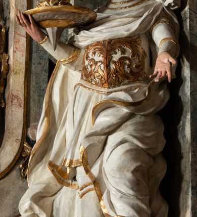 Zdjęcie nr 27: Ołtarz architektoniczny, trójosiowy, z wydzieloną częścią cokołową z bramkami bocznymi, jednokondygnacyjny, ze zwieńczeniem. Mensa drewniana z portatylem z marmuru dębnickiego, umieszczona na drewnianej, prostopadłościennej skrzyni.  Antepedium w kształcie leżącego prostokąta, ujęte profilowaną ramą dekorowaną liśćmi akantu i perełkowaniem. W polu płaskorzeźbione przedstawienie baranka leżącego na krzyżu, na obłoku, ujęte parą liści akantu oraz wicią roślinną z kwiatami i kiściami winogron. 
Na mensie tabernakulum z Tronem Łaski w formie prostopadłościanu na cokole, o wypukłej ściance frontowej, ujęte spływami wolutowymi podtrzymującymi niepełne belkowanie, wyłamane po środku łukiem półkolistym. Na froncie obrotowa nisza ujęta obramieniem w kształcie stojącego prostokąta zamkniętego łukiem nadwieszonym. W niszy ryte przedstawienie kielicha z Eucharystią w glorii, na obłoku, całość w rocaillowej ramie. Powyżej tron wystawienia z cokołem dekorowanym rocaille’em i parą ornamentalnych świeczników; ujęty parą kolumn dźwigających imposty. Podpory dżwigają rocaillowe kabłąki tworzące baldachim, na którego szczycie figurka baranka eucharystycznego leżącego na księdze. Ścianka w formie zbliżonej do stojącego prostokąta ujęta rocaille. Tabernakulum i tron ujęte okazałymi spływami wolutowymi z rocaillowymi uszami oraz na zakończeniach u dołu dwiema rzeźbami klęczących, adorujących aniołów.
Osie nastawy wydzielone w osiach skrajnych parą kolumn o skręconych trzonach i korynckich kapitelach umieszczonych na tle pilastrów, w osiach wewnętrznych parą pilastrów o trzonach w formie wolut z puttami z puttami, podtrzymywaną przez kariatydy w formie okazałych figur uskrzydlonych aniołów. Podpory wspierają pełne belkowanie z profilowanym gzymsem, występujące nad nimi, przerwane nad polem środkowym. W polu środkowym nisza ze sklepieniem kolebkowym wspartym na dwóch filarach i pilastrach. W głębi niszy obraz Św. Bartłomiej w profilowanej ramie w kształcie stojącego prostokąta zamkniętego łukiem odcinkowym. Na podniebiu niszy obraz Męczeństwo św. Bartłomieja w kształcie leżącego prostokąta ujętego niesymetrycznym obramieniem z motywem rocaille’ów. Nisza flankowana z figurami św. Stefana i św. Emeryka umieszczonymi na konsolach, na tle pilastrów. Pola retabulum w osiach skrajnych wklęsłe, między podporami prześwity z figurami św. Piotra i św. Pawła. W partii wysokiego cokołu bramki ujęte od zewnątrz wklęsłymi filarami podtrzymującymi belkowanie, wejścia z drzwiczkami w kształcie stojącego prostokąta zamkniętego łukiem półkolistym z obrazami przedstawiającymi Mojżesza i Aarona. Zwieńczenie w formie aedikuli ujętej pilastrami wspierającymi przerwane belkowanie oraz fantazyjnymi spływami wolutowymi, spływającymi na wewnętrzne podpory, przyjmując formę przerwanego przyczółka, na którym dwa putta i dwie figury siedzących aniołów. W osiach zewnętrznych podpór dwa putta siedzące kanelowanych postumentach i obłokach, prawe przytrzymuje skórę (atrybut św. Bartłomieja). W polu zwieńczenia obraz Św. Anna Samotrzeć w kształcie wydłużonego ośmioboku, w ornamentalnej ramie. Całość zwieńczona rzeźbioną półpostacią Boga Ojca pośród obłoków ukazanego w glorii promienistej. Drzwi do bramek flankowane liściastymi festonami, powyżej tarcze ujęte liśćmi laurowymi, zwieńczone koroną z klejnotem; lewa z klejnotem w postaci łabędzia z napisem: SOLI/ DEO/GLORIA, prawa z postacią ludzką z napisem: SOLI DEO HONOR. Woluty i fryz dekorowane motywami rocaille’ów oraz uskrzydlonymi główkami anielskimi. Osie boczne wypełnione ażurowymi rocaille’ami z gałązkami różanymi oraz ornamentem małżowinowo-chrząstkowym, belkowanie z podwieszonym lambrekinem z chwostami. Skręcone trzony kolumn zdobione wicią z winnej latorośli. Archiwolta nad polem środkowym dekorowana rocaille’em, rocaille’owym kartuszem i lambrekinem z chwostami. Woluty zwieńczenia, pilastry i naroża pola obrazowego dekorowane motywami rocaille’ów. 
Nastawa marmoryzowana w odcieniu jasnoszarym, profile marmoryzowane w kolorze jasnoróżowym, ornamenty i obramienia złocone.