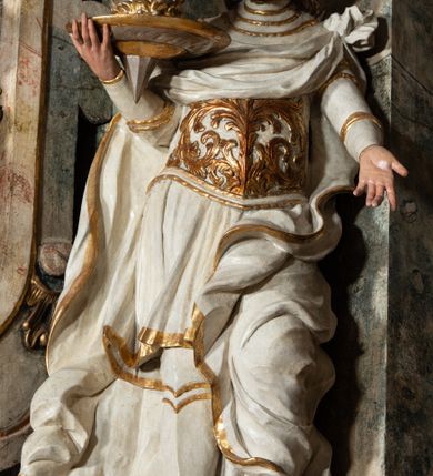 Zdjęcie nr 26: Ołtarz architektoniczny, trójosiowy, z wydzieloną częścią cokołową z bramkami bocznymi, jednokondygnacyjny, ze zwieńczeniem. Mensa drewniana z portatylem z marmuru dębnickiego, umieszczona na drewnianej, prostopadłościennej skrzyni.  Antepedium w kształcie leżącego prostokąta, ujęte profilowaną ramą dekorowaną liśćmi akantu i perełkowaniem. W polu płaskorzeźbione przedstawienie baranka leżącego na krzyżu, na obłoku, ujęte parą liści akantu oraz wicią roślinną z kwiatami i kiściami winogron. 
Na mensie tabernakulum z Tronem Łaski w formie prostopadłościanu na cokole, o wypukłej ściance frontowej, ujęte spływami wolutowymi podtrzymującymi niepełne belkowanie, wyłamane po środku łukiem półkolistym. Na froncie obrotowa nisza ujęta obramieniem w kształcie stojącego prostokąta zamkniętego łukiem nadwieszonym. W niszy ryte przedstawienie kielicha z Eucharystią w glorii, na obłoku, całość w rocaillowej ramie. Powyżej tron wystawienia z cokołem dekorowanym rocaille’em i parą ornamentalnych świeczników; ujęty parą kolumn dźwigających imposty. Podpory dżwigają rocaillowe kabłąki tworzące baldachim, na którego szczycie figurka baranka eucharystycznego leżącego na księdze. Ścianka w formie zbliżonej do stojącego prostokąta ujęta rocaille. Tabernakulum i tron ujęte okazałymi spływami wolutowymi z rocaillowymi uszami oraz na zakończeniach u dołu dwiema rzeźbami klęczących, adorujących aniołów.
Osie nastawy wydzielone w osiach skrajnych parą kolumn o skręconych trzonach i korynckich kapitelach umieszczonych na tle pilastrów, w osiach wewnętrznych parą pilastrów o trzonach w formie wolut z puttami z puttami, podtrzymywaną przez kariatydy w formie okazałych figur uskrzydlonych aniołów. Podpory wspierają pełne belkowanie z profilowanym gzymsem, występujące nad nimi, przerwane nad polem środkowym. W polu środkowym nisza ze sklepieniem kolebkowym wspartym na dwóch filarach i pilastrach. W głębi niszy obraz Św. Bartłomiej w profilowanej ramie w kształcie stojącego prostokąta zamkniętego łukiem odcinkowym. Na podniebiu niszy obraz Męczeństwo św. Bartłomieja w kształcie leżącego prostokąta ujętego niesymetrycznym obramieniem z motywem rocaille’ów. Nisza flankowana z figurami św. Stefana i św. Emeryka umieszczonymi na konsolach, na tle pilastrów. Pola retabulum w osiach skrajnych wklęsłe, między podporami prześwity z figurami św. Piotra i św. Pawła. W partii wysokiego cokołu bramki ujęte od zewnątrz wklęsłymi filarami podtrzymującymi belkowanie, wejścia z drzwiczkami w kształcie stojącego prostokąta zamkniętego łukiem półkolistym z obrazami przedstawiającymi Mojżesza i Aarona. Zwieńczenie w formie aedikuli ujętej pilastrami wspierającymi przerwane belkowanie oraz fantazyjnymi spływami wolutowymi, spływającymi na wewnętrzne podpory, przyjmując formę przerwanego przyczółka, na którym dwa putta i dwie figury siedzących aniołów. W osiach zewnętrznych podpór dwa putta siedzące kanelowanych postumentach i obłokach, prawe przytrzymuje skórę (atrybut św. Bartłomieja). W polu zwieńczenia obraz Św. Anna Samotrzeć w kształcie wydłużonego ośmioboku, w ornamentalnej ramie. Całość zwieńczona rzeźbioną półpostacią Boga Ojca pośród obłoków ukazanego w glorii promienistej. Drzwi do bramek flankowane liściastymi festonami, powyżej tarcze ujęte liśćmi laurowymi, zwieńczone koroną z klejnotem; lewa z klejnotem w postaci łabędzia z napisem: SOLI/ DEO/GLORIA, prawa z postacią ludzką z napisem: SOLI DEO HONOR. Woluty i fryz dekorowane motywami rocaille’ów oraz uskrzydlonymi główkami anielskimi. Osie boczne wypełnione ażurowymi rocaille’ami z gałązkami różanymi oraz ornamentem małżowinowo-chrząstkowym, belkowanie z podwieszonym lambrekinem z chwostami. Skręcone trzony kolumn zdobione wicią z winnej latorośli. Archiwolta nad polem środkowym dekorowana rocaille’em, rocaille’owym kartuszem i lambrekinem z chwostami. Woluty zwieńczenia, pilastry i naroża pola obrazowego dekorowane motywami rocaille’ów. 
Nastawa marmoryzowana w odcieniu jasnoszarym, profile marmoryzowane w kolorze jasnoróżowym, ornamenty i obramienia złocone.