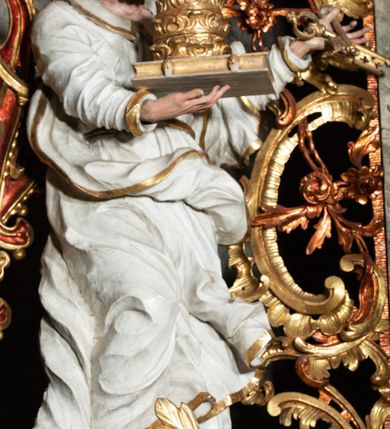 Zdjęcie nr 35: Ołtarz architektoniczny, trójosiowy, z wydzieloną częścią cokołową z bramkami bocznymi, jednokondygnacyjny, ze zwieńczeniem. Mensa drewniana z portatylem z marmuru dębnickiego, umieszczona na drewnianej, prostopadłościennej skrzyni.  Antepedium w kształcie leżącego prostokąta, ujęte profilowaną ramą dekorowaną liśćmi akantu i perełkowaniem. W polu płaskorzeźbione przedstawienie baranka leżącego na krzyżu, na obłoku, ujęte parą liści akantu oraz wicią roślinną z kwiatami i kiściami winogron. 
Na mensie tabernakulum z Tronem Łaski w formie prostopadłościanu na cokole, o wypukłej ściance frontowej, ujęte spływami wolutowymi podtrzymującymi niepełne belkowanie, wyłamane po środku łukiem półkolistym. Na froncie obrotowa nisza ujęta obramieniem w kształcie stojącego prostokąta zamkniętego łukiem nadwieszonym. W niszy ryte przedstawienie kielicha z Eucharystią w glorii, na obłoku, całość w rocaillowej ramie. Powyżej tron wystawienia z cokołem dekorowanym rocaille’em i parą ornamentalnych świeczników; ujęty parą kolumn dźwigających imposty. Podpory dżwigają rocaillowe kabłąki tworzące baldachim, na którego szczycie figurka baranka eucharystycznego leżącego na księdze. Ścianka w formie zbliżonej do stojącego prostokąta ujęta rocaille. Tabernakulum i tron ujęte okazałymi spływami wolutowymi z rocaillowymi uszami oraz na zakończeniach u dołu dwiema rzeźbami klęczących, adorujących aniołów.
Osie nastawy wydzielone w osiach skrajnych parą kolumn o skręconych trzonach i korynckich kapitelach umieszczonych na tle pilastrów, w osiach wewnętrznych parą pilastrów o trzonach w formie wolut z puttami z puttami, podtrzymywaną przez kariatydy w formie okazałych figur uskrzydlonych aniołów. Podpory wspierają pełne belkowanie z profilowanym gzymsem, występujące nad nimi, przerwane nad polem środkowym. W polu środkowym nisza ze sklepieniem kolebkowym wspartym na dwóch filarach i pilastrach. W głębi niszy obraz Św. Bartłomiej w profilowanej ramie w kształcie stojącego prostokąta zamkniętego łukiem odcinkowym. Na podniebiu niszy obraz Męczeństwo św. Bartłomieja w kształcie leżącego prostokąta ujętego niesymetrycznym obramieniem z motywem rocaille’ów. Nisza flankowana z figurami św. Stefana i św. Emeryka umieszczonymi na konsolach, na tle pilastrów. Pola retabulum w osiach skrajnych wklęsłe, między podporami prześwity z figurami św. Piotra i św. Pawła. W partii wysokiego cokołu bramki ujęte od zewnątrz wklęsłymi filarami podtrzymującymi belkowanie, wejścia z drzwiczkami w kształcie stojącego prostokąta zamkniętego łukiem półkolistym z obrazami przedstawiającymi Mojżesza i Aarona. Zwieńczenie w formie aedikuli ujętej pilastrami wspierającymi przerwane belkowanie oraz fantazyjnymi spływami wolutowymi, spływającymi na wewnętrzne podpory, przyjmując formę przerwanego przyczółka, na którym dwa putta i dwie figury siedzących aniołów. W osiach zewnętrznych podpór dwa putta siedzące kanelowanych postumentach i obłokach, prawe przytrzymuje skórę (atrybut św. Bartłomieja). W polu zwieńczenia obraz Św. Anna Samotrzeć w kształcie wydłużonego ośmioboku, w ornamentalnej ramie. Całość zwieńczona rzeźbioną półpostacią Boga Ojca pośród obłoków ukazanego w glorii promienistej. Drzwi do bramek flankowane liściastymi festonami, powyżej tarcze ujęte liśćmi laurowymi, zwieńczone koroną z klejnotem; lewa z klejnotem w postaci łabędzia z napisem: SOLI/ DEO/GLORIA, prawa z postacią ludzką z napisem: SOLI DEO HONOR. Woluty i fryz dekorowane motywami rocaille’ów oraz uskrzydlonymi główkami anielskimi. Osie boczne wypełnione ażurowymi rocaille’ami z gałązkami różanymi oraz ornamentem małżowinowo-chrząstkowym, belkowanie z podwieszonym lambrekinem z chwostami. Skręcone trzony kolumn zdobione wicią z winnej latorośli. Archiwolta nad polem środkowym dekorowana rocaille’em, rocaille’owym kartuszem i lambrekinem z chwostami. Woluty zwieńczenia, pilastry i naroża pola obrazowego dekorowane motywami rocaille’ów. 
Nastawa marmoryzowana w odcieniu jasnoszarym, profile marmoryzowane w kolorze jasnoróżowym, ornamenty i obramienia złocone.