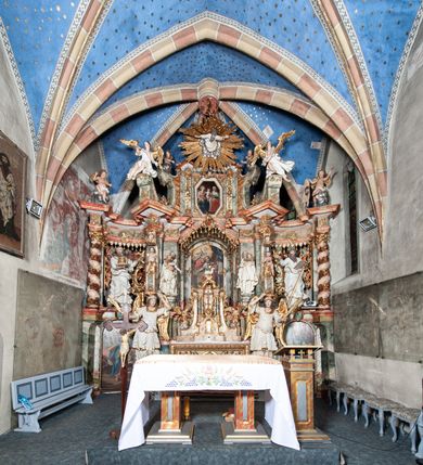 Zdjęcie nr 7: Ołtarz architektoniczny, trójosiowy, z wydzieloną częścią cokołową z bramkami bocznymi, jednokondygnacyjny, ze zwieńczeniem. Mensa drewniana z portatylem z marmuru dębnickiego, umieszczona na drewnianej, prostopadłościennej skrzyni.  Antepedium w kształcie leżącego prostokąta, ujęte profilowaną ramą dekorowaną liśćmi akantu i perełkowaniem. W polu płaskorzeźbione przedstawienie baranka leżącego na krzyżu, na obłoku, ujęte parą liści akantu oraz wicią roślinną z kwiatami i kiściami winogron. 
Na mensie tabernakulum z Tronem Łaski w formie prostopadłościanu na cokole, o wypukłej ściance frontowej, ujęte spływami wolutowymi podtrzymującymi niepełne belkowanie, wyłamane po środku łukiem półkolistym. Na froncie obrotowa nisza ujęta obramieniem w kształcie stojącego prostokąta zamkniętego łukiem nadwieszonym. W niszy ryte przedstawienie kielicha z Eucharystią w glorii, na obłoku, całość w rocaillowej ramie. Powyżej tron wystawienia z cokołem dekorowanym rocaille’em i parą ornamentalnych świeczników; ujęty parą kolumn dźwigających imposty. Podpory dżwigają rocaillowe kabłąki tworzące baldachim, na którego szczycie figurka baranka eucharystycznego leżącego na księdze. Ścianka w formie zbliżonej do stojącego prostokąta ujęta rocaille. Tabernakulum i tron ujęte okazałymi spływami wolutowymi z rocaillowymi uszami oraz na zakończeniach u dołu dwiema rzeźbami klęczących, adorujących aniołów.
Osie nastawy wydzielone w osiach skrajnych parą kolumn o skręconych trzonach i korynckich kapitelach umieszczonych na tle pilastrów, w osiach wewnętrznych parą pilastrów o trzonach w formie wolut z puttami z puttami, podtrzymywaną przez kariatydy w formie okazałych figur uskrzydlonych aniołów. Podpory wspierają pełne belkowanie z profilowanym gzymsem, występujące nad nimi, przerwane nad polem środkowym. W polu środkowym nisza ze sklepieniem kolebkowym wspartym na dwóch filarach i pilastrach. W głębi niszy obraz Św. Bartłomiej w profilowanej ramie w kształcie stojącego prostokąta zamkniętego łukiem odcinkowym. Na podniebiu niszy obraz Męczeństwo św. Bartłomieja w kształcie leżącego prostokąta ujętego niesymetrycznym obramieniem z motywem rocaille’ów. Nisza flankowana z figurami św. Stefana i św. Emeryka umieszczonymi na konsolach, na tle pilastrów. Pola retabulum w osiach skrajnych wklęsłe, między podporami prześwity z figurami św. Piotra i św. Pawła. W partii wysokiego cokołu bramki ujęte od zewnątrz wklęsłymi filarami podtrzymującymi belkowanie, wejścia z drzwiczkami w kształcie stojącego prostokąta zamkniętego łukiem półkolistym z obrazami przedstawiającymi Mojżesza i Aarona. Zwieńczenie w formie aedikuli ujętej pilastrami wspierającymi przerwane belkowanie oraz fantazyjnymi spływami wolutowymi, spływającymi na wewnętrzne podpory, przyjmując formę przerwanego przyczółka, na którym dwa putta i dwie figury siedzących aniołów. W osiach zewnętrznych podpór dwa putta siedzące kanelowanych postumentach i obłokach, prawe przytrzymuje skórę (atrybut św. Bartłomieja). W polu zwieńczenia obraz Św. Anna Samotrzeć w kształcie wydłużonego ośmioboku, w ornamentalnej ramie. Całość zwieńczona rzeźbioną półpostacią Boga Ojca pośród obłoków ukazanego w glorii promienistej. Drzwi do bramek flankowane liściastymi festonami, powyżej tarcze ujęte liśćmi laurowymi, zwieńczone koroną z klejnotem; lewa z klejnotem w postaci łabędzia z napisem: SOLI/ DEO/GLORIA, prawa z postacią ludzką z napisem: SOLI DEO HONOR. Woluty i fryz dekorowane motywami rocaille’ów oraz uskrzydlonymi główkami anielskimi. Osie boczne wypełnione ażurowymi rocaille’ami z gałązkami różanymi oraz ornamentem małżowinowo-chrząstkowym, belkowanie z podwieszonym lambrekinem z chwostami. Skręcone trzony kolumn zdobione wicią z winnej latorośli. Archiwolta nad polem środkowym dekorowana rocaille’em, rocaille’owym kartuszem i lambrekinem z chwostami. Woluty zwieńczenia, pilastry i naroża pola obrazowego dekorowane motywami rocaille’ów. 
Nastawa marmoryzowana w odcieniu jasnoszarym, profile marmoryzowane w kolorze jasnoróżowym, ornamenty i obramienia złocone.