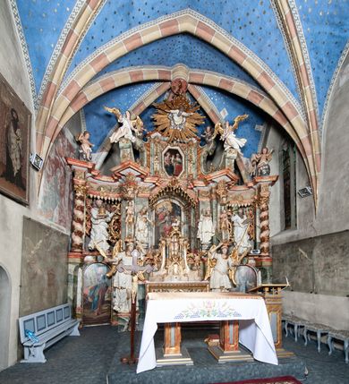 Zdjęcie nr 6: Ołtarz architektoniczny, trójosiowy, z wydzieloną częścią cokołową z bramkami bocznymi, jednokondygnacyjny, ze zwieńczeniem. Mensa drewniana z portatylem z marmuru dębnickiego, umieszczona na drewnianej, prostopadłościennej skrzyni.  Antepedium w kształcie leżącego prostokąta, ujęte profilowaną ramą dekorowaną liśćmi akantu i perełkowaniem. W polu płaskorzeźbione przedstawienie baranka leżącego na krzyżu, na obłoku, ujęte parą liści akantu oraz wicią roślinną z kwiatami i kiściami winogron. 
Na mensie tabernakulum z Tronem Łaski w formie prostopadłościanu na cokole, o wypukłej ściance frontowej, ujęte spływami wolutowymi podtrzymującymi niepełne belkowanie, wyłamane po środku łukiem półkolistym. Na froncie obrotowa nisza ujęta obramieniem w kształcie stojącego prostokąta zamkniętego łukiem nadwieszonym. W niszy ryte przedstawienie kielicha z Eucharystią w glorii, na obłoku, całość w rocaillowej ramie. Powyżej tron wystawienia z cokołem dekorowanym rocaille’em i parą ornamentalnych świeczników; ujęty parą kolumn dźwigających imposty. Podpory dżwigają rocaillowe kabłąki tworzące baldachim, na którego szczycie figurka baranka eucharystycznego leżącego na księdze. Ścianka w formie zbliżonej do stojącego prostokąta ujęta rocaille. Tabernakulum i tron ujęte okazałymi spływami wolutowymi z rocaillowymi uszami oraz na zakończeniach u dołu dwiema rzeźbami klęczących, adorujących aniołów.
Osie nastawy wydzielone w osiach skrajnych parą kolumn o skręconych trzonach i korynckich kapitelach umieszczonych na tle pilastrów, w osiach wewnętrznych parą pilastrów o trzonach w formie wolut z puttami z puttami, podtrzymywaną przez kariatydy w formie okazałych figur uskrzydlonych aniołów. Podpory wspierają pełne belkowanie z profilowanym gzymsem, występujące nad nimi, przerwane nad polem środkowym. W polu środkowym nisza ze sklepieniem kolebkowym wspartym na dwóch filarach i pilastrach. W głębi niszy obraz Św. Bartłomiej w profilowanej ramie w kształcie stojącego prostokąta zamkniętego łukiem odcinkowym. Na podniebiu niszy obraz Męczeństwo św. Bartłomieja w kształcie leżącego prostokąta ujętego niesymetrycznym obramieniem z motywem rocaille’ów. Nisza flankowana z figurami św. Stefana i św. Emeryka umieszczonymi na konsolach, na tle pilastrów. Pola retabulum w osiach skrajnych wklęsłe, między podporami prześwity z figurami św. Piotra i św. Pawła. W partii wysokiego cokołu bramki ujęte od zewnątrz wklęsłymi filarami podtrzymującymi belkowanie, wejścia z drzwiczkami w kształcie stojącego prostokąta zamkniętego łukiem półkolistym z obrazami przedstawiającymi Mojżesza i Aarona. Zwieńczenie w formie aedikuli ujętej pilastrami wspierającymi przerwane belkowanie oraz fantazyjnymi spływami wolutowymi, spływającymi na wewnętrzne podpory, przyjmując formę przerwanego przyczółka, na którym dwa putta i dwie figury siedzących aniołów. W osiach zewnętrznych podpór dwa putta siedzące kanelowanych postumentach i obłokach, prawe przytrzymuje skórę (atrybut św. Bartłomieja). W polu zwieńczenia obraz Św. Anna Samotrzeć w kształcie wydłużonego ośmioboku, w ornamentalnej ramie. Całość zwieńczona rzeźbioną półpostacią Boga Ojca pośród obłoków ukazanego w glorii promienistej. Drzwi do bramek flankowane liściastymi festonami, powyżej tarcze ujęte liśćmi laurowymi, zwieńczone koroną z klejnotem; lewa z klejnotem w postaci łabędzia z napisem: SOLI/ DEO/GLORIA, prawa z postacią ludzką z napisem: SOLI DEO HONOR. Woluty i fryz dekorowane motywami rocaille’ów oraz uskrzydlonymi główkami anielskimi. Osie boczne wypełnione ażurowymi rocaille’ami z gałązkami różanymi oraz ornamentem małżowinowo-chrząstkowym, belkowanie z podwieszonym lambrekinem z chwostami. Skręcone trzony kolumn zdobione wicią z winnej latorośli. Archiwolta nad polem środkowym dekorowana rocaille’em, rocaille’owym kartuszem i lambrekinem z chwostami. Woluty zwieńczenia, pilastry i naroża pola obrazowego dekorowane motywami rocaille’ów. 
Nastawa marmoryzowana w odcieniu jasnoszarym, profile marmoryzowane w kolorze jasnoróżowym, ornamenty i obramienia złocone.