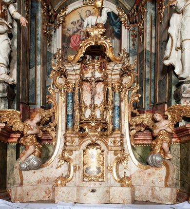 Zdjęcie nr 17: Ołtarz architektoniczny, trójosiowy, z wydzieloną częścią cokołową z bramkami bocznymi, jednokondygnacyjny, ze zwieńczeniem. Mensa drewniana z portatylem z marmuru dębnickiego, umieszczona na drewnianej, prostopadłościennej skrzyni.  Antepedium w kształcie leżącego prostokąta, ujęte profilowaną ramą dekorowaną liśćmi akantu i perełkowaniem. W polu płaskorzeźbione przedstawienie baranka leżącego na krzyżu, na obłoku, ujęte parą liści akantu oraz wicią roślinną z kwiatami i kiściami winogron. 
Na mensie tabernakulum z Tronem Łaski w formie prostopadłościanu na cokole, o wypukłej ściance frontowej, ujęte spływami wolutowymi podtrzymującymi niepełne belkowanie, wyłamane po środku łukiem półkolistym. Na froncie obrotowa nisza ujęta obramieniem w kształcie stojącego prostokąta zamkniętego łukiem nadwieszonym. W niszy ryte przedstawienie kielicha z Eucharystią w glorii, na obłoku, całość w rocaillowej ramie. Powyżej tron wystawienia z cokołem dekorowanym rocaille’em i parą ornamentalnych świeczników; ujęty parą kolumn dźwigających imposty. Podpory dżwigają rocaillowe kabłąki tworzące baldachim, na którego szczycie figurka baranka eucharystycznego leżącego na księdze. Ścianka w formie zbliżonej do stojącego prostokąta ujęta rocaille. Tabernakulum i tron ujęte okazałymi spływami wolutowymi z rocaillowymi uszami oraz na zakończeniach u dołu dwiema rzeźbami klęczących, adorujących aniołów.
Osie nastawy wydzielone w osiach skrajnych parą kolumn o skręconych trzonach i korynckich kapitelach umieszczonych na tle pilastrów, w osiach wewnętrznych parą pilastrów o trzonach w formie wolut z puttami z puttami, podtrzymywaną przez kariatydy w formie okazałych figur uskrzydlonych aniołów. Podpory wspierają pełne belkowanie z profilowanym gzymsem, występujące nad nimi, przerwane nad polem środkowym. W polu środkowym nisza ze sklepieniem kolebkowym wspartym na dwóch filarach i pilastrach. W głębi niszy obraz Św. Bartłomiej w profilowanej ramie w kształcie stojącego prostokąta zamkniętego łukiem odcinkowym. Na podniebiu niszy obraz Męczeństwo św. Bartłomieja w kształcie leżącego prostokąta ujętego niesymetrycznym obramieniem z motywem rocaille’ów. Nisza flankowana z figurami św. Stefana i św. Emeryka umieszczonymi na konsolach, na tle pilastrów. Pola retabulum w osiach skrajnych wklęsłe, między podporami prześwity z figurami św. Piotra i św. Pawła. W partii wysokiego cokołu bramki ujęte od zewnątrz wklęsłymi filarami podtrzymującymi belkowanie, wejścia z drzwiczkami w kształcie stojącego prostokąta zamkniętego łukiem półkolistym z obrazami przedstawiającymi Mojżesza i Aarona. Zwieńczenie w formie aedikuli ujętej pilastrami wspierającymi przerwane belkowanie oraz fantazyjnymi spływami wolutowymi, spływającymi na wewnętrzne podpory, przyjmując formę przerwanego przyczółka, na którym dwa putta i dwie figury siedzących aniołów. W osiach zewnętrznych podpór dwa putta siedzące kanelowanych postumentach i obłokach, prawe przytrzymuje skórę (atrybut św. Bartłomieja). W polu zwieńczenia obraz Św. Anna Samotrzeć w kształcie wydłużonego ośmioboku, w ornamentalnej ramie. Całość zwieńczona rzeźbioną półpostacią Boga Ojca pośród obłoków ukazanego w glorii promienistej. Drzwi do bramek flankowane liściastymi festonami, powyżej tarcze ujęte liśćmi laurowymi, zwieńczone koroną z klejnotem; lewa z klejnotem w postaci łabędzia z napisem: SOLI/ DEO/GLORIA, prawa z postacią ludzką z napisem: SOLI DEO HONOR. Woluty i fryz dekorowane motywami rocaille’ów oraz uskrzydlonymi główkami anielskimi. Osie boczne wypełnione ażurowymi rocaille’ami z gałązkami różanymi oraz ornamentem małżowinowo-chrząstkowym, belkowanie z podwieszonym lambrekinem z chwostami. Skręcone trzony kolumn zdobione wicią z winnej latorośli. Archiwolta nad polem środkowym dekorowana rocaille’em, rocaille’owym kartuszem i lambrekinem z chwostami. Woluty zwieńczenia, pilastry i naroża pola obrazowego dekorowane motywami rocaille’ów. 
Nastawa marmoryzowana w odcieniu jasnoszarym, profile marmoryzowane w kolorze jasnoróżowym, ornamenty i obramienia złocone.