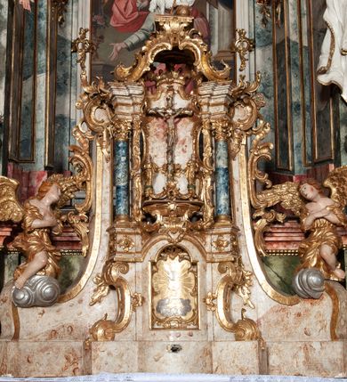 Zdjęcie nr 16: Ołtarz architektoniczny, trójosiowy, z wydzieloną częścią cokołową z bramkami bocznymi, jednokondygnacyjny, ze zwieńczeniem. Mensa drewniana z portatylem z marmuru dębnickiego, umieszczona na drewnianej, prostopadłościennej skrzyni.  Antepedium w kształcie leżącego prostokąta, ujęte profilowaną ramą dekorowaną liśćmi akantu i perełkowaniem. W polu płaskorzeźbione przedstawienie baranka leżącego na krzyżu, na obłoku, ujęte parą liści akantu oraz wicią roślinną z kwiatami i kiściami winogron. 
Na mensie tabernakulum z Tronem Łaski w formie prostopadłościanu na cokole, o wypukłej ściance frontowej, ujęte spływami wolutowymi podtrzymującymi niepełne belkowanie, wyłamane po środku łukiem półkolistym. Na froncie obrotowa nisza ujęta obramieniem w kształcie stojącego prostokąta zamkniętego łukiem nadwieszonym. W niszy ryte przedstawienie kielicha z Eucharystią w glorii, na obłoku, całość w rocaillowej ramie. Powyżej tron wystawienia z cokołem dekorowanym rocaille’em i parą ornamentalnych świeczników; ujęty parą kolumn dźwigających imposty. Podpory dżwigają rocaillowe kabłąki tworzące baldachim, na którego szczycie figurka baranka eucharystycznego leżącego na księdze. Ścianka w formie zbliżonej do stojącego prostokąta ujęta rocaille. Tabernakulum i tron ujęte okazałymi spływami wolutowymi z rocaillowymi uszami oraz na zakończeniach u dołu dwiema rzeźbami klęczących, adorujących aniołów.
Osie nastawy wydzielone w osiach skrajnych parą kolumn o skręconych trzonach i korynckich kapitelach umieszczonych na tle pilastrów, w osiach wewnętrznych parą pilastrów o trzonach w formie wolut z puttami z puttami, podtrzymywaną przez kariatydy w formie okazałych figur uskrzydlonych aniołów. Podpory wspierają pełne belkowanie z profilowanym gzymsem, występujące nad nimi, przerwane nad polem środkowym. W polu środkowym nisza ze sklepieniem kolebkowym wspartym na dwóch filarach i pilastrach. W głębi niszy obraz Św. Bartłomiej w profilowanej ramie w kształcie stojącego prostokąta zamkniętego łukiem odcinkowym. Na podniebiu niszy obraz Męczeństwo św. Bartłomieja w kształcie leżącego prostokąta ujętego niesymetrycznym obramieniem z motywem rocaille’ów. Nisza flankowana z figurami św. Stefana i św. Emeryka umieszczonymi na konsolach, na tle pilastrów. Pola retabulum w osiach skrajnych wklęsłe, między podporami prześwity z figurami św. Piotra i św. Pawła. W partii wysokiego cokołu bramki ujęte od zewnątrz wklęsłymi filarami podtrzymującymi belkowanie, wejścia z drzwiczkami w kształcie stojącego prostokąta zamkniętego łukiem półkolistym z obrazami przedstawiającymi Mojżesza i Aarona. Zwieńczenie w formie aedikuli ujętej pilastrami wspierającymi przerwane belkowanie oraz fantazyjnymi spływami wolutowymi, spływającymi na wewnętrzne podpory, przyjmując formę przerwanego przyczółka, na którym dwa putta i dwie figury siedzących aniołów. W osiach zewnętrznych podpór dwa putta siedzące kanelowanych postumentach i obłokach, prawe przytrzymuje skórę (atrybut św. Bartłomieja). W polu zwieńczenia obraz Św. Anna Samotrzeć w kształcie wydłużonego ośmioboku, w ornamentalnej ramie. Całość zwieńczona rzeźbioną półpostacią Boga Ojca pośród obłoków ukazanego w glorii promienistej. Drzwi do bramek flankowane liściastymi festonami, powyżej tarcze ujęte liśćmi laurowymi, zwieńczone koroną z klejnotem; lewa z klejnotem w postaci łabędzia z napisem: SOLI/ DEO/GLORIA, prawa z postacią ludzką z napisem: SOLI DEO HONOR. Woluty i fryz dekorowane motywami rocaille’ów oraz uskrzydlonymi główkami anielskimi. Osie boczne wypełnione ażurowymi rocaille’ami z gałązkami różanymi oraz ornamentem małżowinowo-chrząstkowym, belkowanie z podwieszonym lambrekinem z chwostami. Skręcone trzony kolumn zdobione wicią z winnej latorośli. Archiwolta nad polem środkowym dekorowana rocaille’em, rocaille’owym kartuszem i lambrekinem z chwostami. Woluty zwieńczenia, pilastry i naroża pola obrazowego dekorowane motywami rocaille’ów. 
Nastawa marmoryzowana w odcieniu jasnoszarym, profile marmoryzowane w kolorze jasnoróżowym, ornamenty i obramienia złocone.