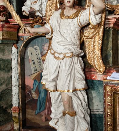 Zdjęcie nr 49: Ołtarz architektoniczny, trójosiowy, z wydzieloną częścią cokołową z bramkami bocznymi, jednokondygnacyjny, ze zwieńczeniem. Mensa drewniana z portatylem z marmuru dębnickiego, umieszczona na drewnianej, prostopadłościennej skrzyni.  Antepedium w kształcie leżącego prostokąta, ujęte profilowaną ramą dekorowaną liśćmi akantu i perełkowaniem. W polu płaskorzeźbione przedstawienie baranka leżącego na krzyżu, na obłoku, ujęte parą liści akantu oraz wicią roślinną z kwiatami i kiściami winogron. 
Na mensie tabernakulum z Tronem Łaski w formie prostopadłościanu na cokole, o wypukłej ściance frontowej, ujęte spływami wolutowymi podtrzymującymi niepełne belkowanie, wyłamane po środku łukiem półkolistym. Na froncie obrotowa nisza ujęta obramieniem w kształcie stojącego prostokąta zamkniętego łukiem nadwieszonym. W niszy ryte przedstawienie kielicha z Eucharystią w glorii, na obłoku, całość w rocaillowej ramie. Powyżej tron wystawienia z cokołem dekorowanym rocaille’em i parą ornamentalnych świeczników; ujęty parą kolumn dźwigających imposty. Podpory dżwigają rocaillowe kabłąki tworzące baldachim, na którego szczycie figurka baranka eucharystycznego leżącego na księdze. Ścianka w formie zbliżonej do stojącego prostokąta ujęta rocaille. Tabernakulum i tron ujęte okazałymi spływami wolutowymi z rocaillowymi uszami oraz na zakończeniach u dołu dwiema rzeźbami klęczących, adorujących aniołów.
Osie nastawy wydzielone w osiach skrajnych parą kolumn o skręconych trzonach i korynckich kapitelach umieszczonych na tle pilastrów, w osiach wewnętrznych parą pilastrów o trzonach w formie wolut z puttami z puttami, podtrzymywaną przez kariatydy w formie okazałych figur uskrzydlonych aniołów. Podpory wspierają pełne belkowanie z profilowanym gzymsem, występujące nad nimi, przerwane nad polem środkowym. W polu środkowym nisza ze sklepieniem kolebkowym wspartym na dwóch filarach i pilastrach. W głębi niszy obraz Św. Bartłomiej w profilowanej ramie w kształcie stojącego prostokąta zamkniętego łukiem odcinkowym. Na podniebiu niszy obraz Męczeństwo św. Bartłomieja w kształcie leżącego prostokąta ujętego niesymetrycznym obramieniem z motywem rocaille’ów. Nisza flankowana z figurami św. Stefana i św. Emeryka umieszczonymi na konsolach, na tle pilastrów. Pola retabulum w osiach skrajnych wklęsłe, między podporami prześwity z figurami św. Piotra i św. Pawła. W partii wysokiego cokołu bramki ujęte od zewnątrz wklęsłymi filarami podtrzymującymi belkowanie, wejścia z drzwiczkami w kształcie stojącego prostokąta zamkniętego łukiem półkolistym z obrazami przedstawiającymi Mojżesza i Aarona. Zwieńczenie w formie aedikuli ujętej pilastrami wspierającymi przerwane belkowanie oraz fantazyjnymi spływami wolutowymi, spływającymi na wewnętrzne podpory, przyjmując formę przerwanego przyczółka, na którym dwa putta i dwie figury siedzących aniołów. W osiach zewnętrznych podpór dwa putta siedzące kanelowanych postumentach i obłokach, prawe przytrzymuje skórę (atrybut św. Bartłomieja). W polu zwieńczenia obraz Św. Anna Samotrzeć w kształcie wydłużonego ośmioboku, w ornamentalnej ramie. Całość zwieńczona rzeźbioną półpostacią Boga Ojca pośród obłoków ukazanego w glorii promienistej. Drzwi do bramek flankowane liściastymi festonami, powyżej tarcze ujęte liśćmi laurowymi, zwieńczone koroną z klejnotem; lewa z klejnotem w postaci łabędzia z napisem: SOLI/ DEO/GLORIA, prawa z postacią ludzką z napisem: SOLI DEO HONOR. Woluty i fryz dekorowane motywami rocaille’ów oraz uskrzydlonymi główkami anielskimi. Osie boczne wypełnione ażurowymi rocaille’ami z gałązkami różanymi oraz ornamentem małżowinowo-chrząstkowym, belkowanie z podwieszonym lambrekinem z chwostami. Skręcone trzony kolumn zdobione wicią z winnej latorośli. Archiwolta nad polem środkowym dekorowana rocaille’em, rocaille’owym kartuszem i lambrekinem z chwostami. Woluty zwieńczenia, pilastry i naroża pola obrazowego dekorowane motywami rocaille’ów. 
Nastawa marmoryzowana w odcieniu jasnoszarym, profile marmoryzowane w kolorze jasnoróżowym, ornamenty i obramienia złocone.