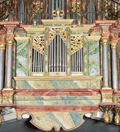 Zdjęcie nr 3: Prospekt architektoniczny, pięcioosiowy, na cokole. Oś środkowa i skrajne wyznaczone ryzalitami. Ryzalit środkowy wyższy od skrajnych, wieloboczny, ryzality skrajne dwuboczne (kątowe); we wszystkich osiach prześwity wypełnione piszczałkami; zwieńczone belkowaniem, podtrzymywane przez wsporniki. Prospekt ujęty uszami utworzonymi z motywów suchego akantu i karbowanej wstęgi. W zwieńczeniu ryzalitu środkowego osiem kabłąków dekorowanych perełkowaniem podtrzymujących rzeźbionego ptaka (kukułka). Ryzality zewnętrzne zwieńczone ażurowymi, trójkątnymi szczytami utworzonymi z motywów roślinnych. Pomiędzy ryzalitami oraz w górnych częściach prześwitów ornament małżowinowy, w dolnej fryz palmetowy. Struktura marmoryzowana na niebiesko, profile i gzyms na czerwono, ornamentyka złocona. Parapet chóru muzycznego, z wypukłą, półokrągłą częścią środkową, wspartą parze filarów stojących na wysokich graniastosłupowych cokołach, o kanelowanych trzonach i akantowych kapitelach, podzielony na siedem pól wydzielonych korynckimi kolumienkami, o gładkich, srebrzonych trzonach, złoconych bazach o kapitelach, stojącymi na konsolkach z podwieszeniem w kształcie szyszki. Kolumienki wspierają wyłamujący się nad nimi gzyms parapetu. Pola w kształcie leżącego prostokąta, ujęte fantazyjnie ukształtowanymi fartuchami, zdobione na przemian obrazami w kształcie ośmioboku lub leżącego owalu opisanego na kwadracie. Obrazy ujęte dekoracyjnymi, profilowanymi ramami. W górnej partii każdego przedstawienia wstęga z napisem: (od lewej): PARABOLA DE PARANDIS PRO TURI ET BELLO Sublibus. Luc. 14 V 2: PARABOLA EPULONIS ET LAZARI Luc. 16 V. 19; PARABOLA PHARISAEI ET PUBLICANI Luc. 18. V. 9; W polu środkowym mały trójosiowy pozytyw. PARABOLA DECEM VIRGINUM MATTH. 23. V. 1; CHRISTUS TRANSFIGURATUR MATTH. 17. V. 1; PROHIBET NIMIAM PROVICTU CURAM MATTH. 6. 25. Struktura marmoryzowana w odcieniach zieleni, beżów i czerwieni; ornamentyka i detal architektoniczny złocony i srebrzony. 