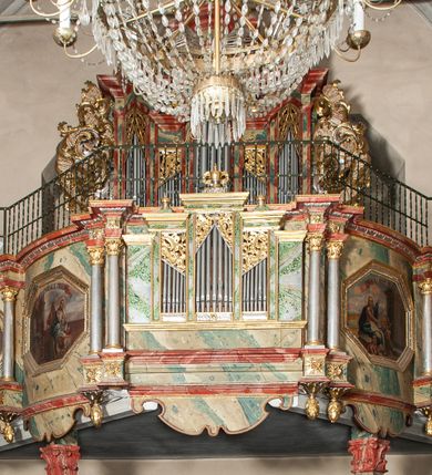 Zdjęcie nr 1: Prospekt architektoniczny, pięcioosiowy, na cokole. Oś środkowa i skrajne wyznaczone ryzalitami. Ryzalit środkowy wyższy od skrajnych, wieloboczny, ryzality skrajne dwuboczne (kątowe); we wszystkich osiach prześwity wypełnione piszczałkami; zwieńczone belkowaniem, podtrzymywane przez wsporniki. Prospekt ujęty uszami utworzonymi z motywów suchego akantu i karbowanej wstęgi. W zwieńczeniu ryzalitu środkowego osiem kabłąków dekorowanych perełkowaniem podtrzymujących rzeźbionego ptaka (kukułka). Ryzality zewnętrzne zwieńczone ażurowymi, trójkątnymi szczytami utworzonymi z motywów roślinnych. Pomiędzy ryzalitami oraz w górnych częściach prześwitów ornament małżowinowy, w dolnej fryz palmetowy. Struktura marmoryzowana na niebiesko, profile i gzyms na czerwono, ornamentyka złocona. Parapet chóru muzycznego, z wypukłą, półokrągłą częścią środkową, wspartą parze filarów stojących na wysokich graniastosłupowych cokołach, o kanelowanych trzonach i akantowych kapitelach, podzielony na siedem pól wydzielonych korynckimi kolumienkami, o gładkich, srebrzonych trzonach, złoconych bazach o kapitelach, stojącymi na konsolkach z podwieszeniem w kształcie szyszki. Kolumienki wspierają wyłamujący się nad nimi gzyms parapetu. Pola w kształcie leżącego prostokąta, ujęte fantazyjnie ukształtowanymi fartuchami, zdobione na przemian obrazami w kształcie ośmioboku lub leżącego owalu opisanego na kwadracie. Obrazy ujęte dekoracyjnymi, profilowanymi ramami. W górnej partii każdego przedstawienia wstęga z napisem: (od lewej): PARABOLA DE PARANDIS PRO TURI ET BELLO Sublibus. Luc. 14 V 2: PARABOLA EPULONIS ET LAZARI Luc. 16 V. 19; PARABOLA PHARISAEI ET PUBLICANI Luc. 18. V. 9; W polu środkowym mały trójosiowy pozytyw. PARABOLA DECEM VIRGINUM MATTH. 23. V. 1; CHRISTUS TRANSFIGURATUR MATTH. 17. V. 1; PROHIBET NIMIAM PROVICTU CURAM MATTH. 6. 25. Struktura marmoryzowana w odcieniach zieleni, beżów i czerwieni; ornamentyka i detal architektoniczny złocony i srebrzony. 