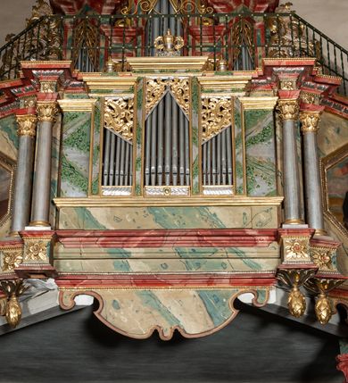 Zdjęcie nr 2: Prospekt architektoniczny, pięcioosiowy, na cokole. Oś środkowa i skrajne wyznaczone ryzalitami. Ryzalit środkowy wyższy od skrajnych, wieloboczny, ryzality skrajne dwuboczne (kątowe); we wszystkich osiach prześwity wypełnione piszczałkami; zwieńczone belkowaniem, podtrzymywane przez wsporniki. Prospekt ujęty uszami utworzonymi z motywów suchego akantu i karbowanej wstęgi. W zwieńczeniu ryzalitu środkowego osiem kabłąków dekorowanych perełkowaniem podtrzymujących rzeźbionego ptaka (kukułka). Ryzality zewnętrzne zwieńczone ażurowymi, trójkątnymi szczytami utworzonymi z motywów roślinnych. Pomiędzy ryzalitami oraz w górnych częściach prześwitów ornament małżowinowy, w dolnej fryz palmetowy. Struktura marmoryzowana na niebiesko, profile i gzyms na czerwono, ornamentyka złocona. Parapet chóru muzycznego, z wypukłą, półokrągłą częścią środkową, wspartą parze filarów stojących na wysokich graniastosłupowych cokołach, o kanelowanych trzonach i akantowych kapitelach, podzielony na siedem pól wydzielonych korynckimi kolumienkami, o gładkich, srebrzonych trzonach, złoconych bazach o kapitelach, stojącymi na konsolkach z podwieszeniem w kształcie szyszki. Kolumienki wspierają wyłamujący się nad nimi gzyms parapetu. Pola w kształcie leżącego prostokąta, ujęte fantazyjnie ukształtowanymi fartuchami, zdobione na przemian obrazami w kształcie ośmioboku lub leżącego owalu opisanego na kwadracie. Obrazy ujęte dekoracyjnymi, profilowanymi ramami. W górnej partii każdego przedstawienia wstęga z napisem: (od lewej): PARABOLA DE PARANDIS PRO TURI ET BELLO Sublibus. Luc. 14 V 2: PARABOLA EPULONIS ET LAZARI Luc. 16 V. 19; PARABOLA PHARISAEI ET PUBLICANI Luc. 18. V. 9; W polu środkowym mały trójosiowy pozytyw. PARABOLA DECEM VIRGINUM MATTH. 23. V. 1; CHRISTUS TRANSFIGURATUR MATTH. 17. V. 1; PROHIBET NIMIAM PROVICTU CURAM MATTH. 6. 25. Struktura marmoryzowana w odcieniach zieleni, beżów i czerwieni; ornamentyka i detal architektoniczny złocony i srebrzony. 