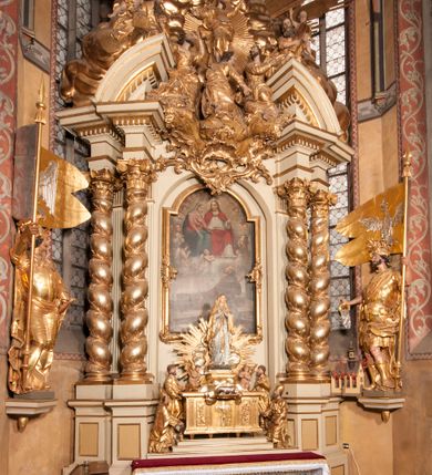 Zdjęcie nr 28: Nastawa jednoosiowa, jednokondygnacyjna ze zwieńczeniem, na wysokim dwustrefowym cokole z wyodrębnionymi czterema prostopadłościanami. Ołtarz kamienny, prostopadłościenny, z kamienną mensą. Antepedium ujęte srebrzoną opaską z napisem „kości ich niech wypuszczą pędu ze swego miejsca, aby byli wsławieni przez swoich synów”, pole podzielone na cztery części ornamentem roślinnym, każda wypełniona złoconą wicią kwiatową i czterema okrągłymi prześwitami. Na ołtarzu ustawione tabernakulum w formie Arki Przymierza. Nastawa ujęta dwiema parami pilastrów i dwiema parami kolumn o kręconych trzonach ustawionych pod kątem na zewnątrz, dźwigającymi pełne belkowanie z kostkowaniem, wyłamane na ich osi, przerwane nad polem środkowym oraz fragmenty przerwanego, półkolistego przyczółka. W polu środkowym wnęka w formie stojącego prostokąta zamkniętego łukiem półkolistym, w niej mniejszy obraz w kształcie stojącego prostokąta zamkniętego łukiem półkolistym nadwieszonym z przedstawieniem Wizji św. Franciszka, ujęty ramą dekorowaną rocaille&#039;em. Zwieńczenie w formie grupy rzeźbiarskiej Przemienienia Pańskiego na tle obłoków i w glorii promienistej. 
W partii belkowania nad polem głównym, na rzeźbionej skale podtrzymywanej przez rocaille klęczące postacie św. Piotra, św. Jana i św. Jakuba w dynamicznych pozach. Nad nimi unosi się Chrystus w otoku z obłoków i glorii, flankowany przez figury Mojżesza i Eliasza klęczące na przerwanym przyczółku, za nimi nadlatujące putta na obłokach. Po lewej stronie Piotr klęczący frontalnie z uniesionymi rękami. Obok niego klęczący św. Jan zwrócony profilem w lewo, z rękami wyciągniętymi w przód, o młodzieńczych rysach i długich, kręconych włosach. Święty Jakub po prawej stronie, w pozycji siedzącej, zwrócony w trzech czwartych w prawo z rękami wyciągniętymi w lewo. Wszyscy ubrani w przylegające do ciała tuniki oraz płaszcze przepasane w partii nóg. Nad nimi Chrystus w pozycji stojącej, frontalnie, w kontrapoście, z uniesioną głową i rękami rozłożonymi na boki, ubrany jest w tunikę oraz płaszcz przewiązany przez lewe ramię i wokół nóg. Na lewym fragmencie przyczółka klęczy Eliasz, zwrócony w trzech czwartych w lewo, pochylony, prawą ręką przytrzymuje na boku księgę, lewą wyciąga w przód. Ubrany jest w tunikę oraz rozwiany płaszcz. Na prawym fragmencie przyczółka klęczy Mojżesz, zwrócony w trzech czwartych w prawo, z pochylona głową, obiema rękami trzyma tablice przykazań. Ubrany jest w tunikę o krótkich rękawach oraz płaszcz zawinięty na lewym ramieniu i opadający na nogi. Obaj prorocy ukazani jako starcy. Nastawa flankowana dwoma wspornikami z figurami św. Wacława i św. Floriana. Struktura polichromowana w kolorze beżowym z żółtymi detalami oraz złoconymi profilami, ornamentyką i zwieńczeniem. Figury w partii ciała polichromowane naturalistycznie z brązowymi włosami i brodami oraz złoconymi szatami i atrybutami. 

