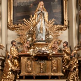 Zdjęcie nr 1: Tabernakulum w formie Arki Przymierza, adorowanej przez dwie klęczące figury starotestamentowego arcykapłana i biskupa oraz dwie stojące postacie lewitów oraz klęczące aniołów, zwieńczonej glorią promienistą z okiem Opatrzności. Wsparte na ośmiu cylindrycznych nóżkach prostopadłościenne tabernakulum jest od frontu podzielone na trzy pola wyznaczone kanelowanymi listwami. Pole środkowe stanowią drzwiczki z rytymi cyframi Dekalogu z ukrzyżowanym Chrystusem, pola boczne z wiązką pałek wodnych z jednej i winnym gronem z drugiej strony. Na skłębionych chmurach z podestem leży Baranek (obecnie w jego miejscu znajduje się figura Matki Boskiej). W tle znacznej wielkości gloria promienista z wieńcem chmur i umieszczonym w nim okiem Opatrzności oraz dwie uskrzydlone główki, nad którymi papieska tiara ze skrzyżowanymi kluczami. Po bokach Baranka dwa klęczące, uskrzydlone nagie aniołki. Obok tabernakulum dwie klęczące postacie z kadzielnicami w rękach. Postać po lewej w stroju biskupim, w albie, stule i kapie oraz infule z dwiema taśmami. Postać po prawej stronie jest ubrany w tradycyjny starotestamentowy strój – efod czyli tunikę i płaszcz wierzchni przepasany na biodrach, pektorał na piersi i zawój na głowie. Za nimi dwaj stojący lewici w tunikach przepasanych na biodrach i wysokich butach. Adorujące Baranka anioły w pozycji klęczącej z rękami skrzyżowanymi na piersi, ze skrzydłami na plecach i nogami w połowie utworzonymi ze skrzydeł.  


