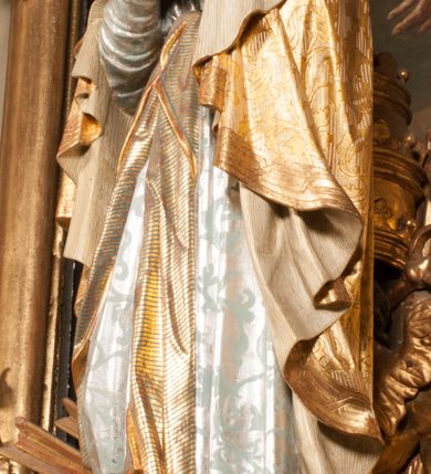 Zdjęcie nr 96: Tabernakulum w formie Arki Przymierza, adorowanej przez dwie klęczące figury starotestamentowego arcykapłana i biskupa oraz dwie stojące postacie lewitów oraz klęczące aniołów, zwieńczonej glorią promienistą z okiem Opatrzności. Wsparte na ośmiu cylindrycznych nóżkach prostopadłościenne tabernakulum jest od frontu podzielone na trzy pola wyznaczone kanelowanymi listwami. Pole środkowe stanowią drzwiczki z rytymi cyframi Dekalogu z ukrzyżowanym Chrystusem, pola boczne z wiązką pałek wodnych z jednej i winnym gronem z drugiej strony. Na skłębionych chmurach z podestem leży Baranek (obecnie w jego miejscu znajduje się figura Matki Boskiej). W tle znacznej wielkości gloria promienista z wieńcem chmur i umieszczonym w nim okiem Opatrzności oraz dwie uskrzydlone główki, nad którymi papieska tiara ze skrzyżowanymi kluczami. Po bokach Baranka dwa klęczące, uskrzydlone nagie aniołki. Obok tabernakulum dwie klęczące postacie z kadzielnicami w rękach. Postać po lewej w stroju biskupim, w albie, stule i kapie oraz infule z dwiema taśmami. Postać po prawej stronie jest ubrany w tradycyjny starotestamentowy strój – efod czyli tunikę i płaszcz wierzchni przepasany na biodrach, pektorał na piersi i zawój na głowie. Za nimi dwaj stojący lewici w tunikach przepasanych na biodrach i wysokich butach. Adorujące Baranka anioły w pozycji klęczącej z rękami skrzyżowanymi na piersi, ze skrzydłami na plecach i nogami w połowie utworzonymi ze skrzydeł.  


