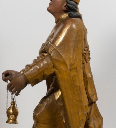 Zdjęcie nr 5: Tabernakulum w formie Arki Przymierza, adorowanej przez dwie klęczące figury starotestamentowego arcykapłana i biskupa oraz dwie stojące postacie lewitów oraz klęczące aniołów, zwieńczonej glorią promienistą z okiem Opatrzności. Wsparte na ośmiu cylindrycznych nóżkach prostopadłościenne tabernakulum jest od frontu podzielone na trzy pola wyznaczone kanelowanymi listwami. Pole środkowe stanowią drzwiczki z rytymi cyframi Dekalogu z ukrzyżowanym Chrystusem, pola boczne z wiązką pałek wodnych z jednej i winnym gronem z drugiej strony. Na skłębionych chmurach z podestem leży Baranek (obecnie w jego miejscu znajduje się figura Matki Boskiej). W tle znacznej wielkości gloria promienista z wieńcem chmur i umieszczonym w nim okiem Opatrzności oraz dwie uskrzydlone główki, nad którymi papieska tiara ze skrzyżowanymi kluczami. Po bokach Baranka dwa klęczące, uskrzydlone nagie aniołki. Obok tabernakulum dwie klęczące postacie z kadzielnicami w rękach. Postać po lewej w stroju biskupim, w albie, stule i kapie oraz infule z dwiema taśmami. Postać po prawej stronie jest ubrany w tradycyjny starotestamentowy strój – efod czyli tunikę i płaszcz wierzchni przepasany na biodrach, pektorał na piersi i zawój na głowie. Za nimi dwaj stojący lewici w tunikach przepasanych na biodrach i wysokich butach. Adorujące Baranka anioły w pozycji klęczącej z rękami skrzyżowanymi na piersi, ze skrzydłami na plecach i nogami w połowie utworzonymi ze skrzydeł.  


