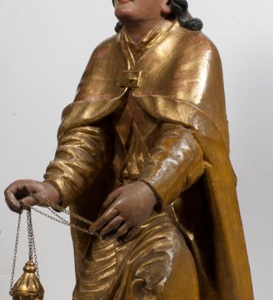 Zdjęcie nr 4: Tabernakulum w formie Arki Przymierza, adorowanej przez dwie klęczące figury starotestamentowego arcykapłana i biskupa oraz dwie stojące postacie lewitów oraz klęczące aniołów, zwieńczonej glorią promienistą z okiem Opatrzności. Wsparte na ośmiu cylindrycznych nóżkach prostopadłościenne tabernakulum jest od frontu podzielone na trzy pola wyznaczone kanelowanymi listwami. Pole środkowe stanowią drzwiczki z rytymi cyframi Dekalogu z ukrzyżowanym Chrystusem, pola boczne z wiązką pałek wodnych z jednej i winnym gronem z drugiej strony. Na skłębionych chmurach z podestem leży Baranek (obecnie w jego miejscu znajduje się figura Matki Boskiej). W tle znacznej wielkości gloria promienista z wieńcem chmur i umieszczonym w nim okiem Opatrzności oraz dwie uskrzydlone główki, nad którymi papieska tiara ze skrzyżowanymi kluczami. Po bokach Baranka dwa klęczące, uskrzydlone nagie aniołki. Obok tabernakulum dwie klęczące postacie z kadzielnicami w rękach. Postać po lewej w stroju biskupim, w albie, stule i kapie oraz infule z dwiema taśmami. Postać po prawej stronie jest ubrany w tradycyjny starotestamentowy strój – efod czyli tunikę i płaszcz wierzchni przepasany na biodrach, pektorał na piersi i zawój na głowie. Za nimi dwaj stojący lewici w tunikach przepasanych na biodrach i wysokich butach. Adorujące Baranka anioły w pozycji klęczącej z rękami skrzyżowanymi na piersi, ze skrzydłami na plecach i nogami w połowie utworzonymi ze skrzydeł.  


