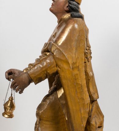 Zdjęcie nr 7: Tabernakulum w formie Arki Przymierza, adorowanej przez dwie klęczące figury starotestamentowego arcykapłana i biskupa oraz dwie stojące postacie lewitów oraz klęczące aniołów, zwieńczonej glorią promienistą z okiem Opatrzności. Wsparte na ośmiu cylindrycznych nóżkach prostopadłościenne tabernakulum jest od frontu podzielone na trzy pola wyznaczone kanelowanymi listwami. Pole środkowe stanowią drzwiczki z rytymi cyframi Dekalogu z ukrzyżowanym Chrystusem, pola boczne z wiązką pałek wodnych z jednej i winnym gronem z drugiej strony. Na skłębionych chmurach z podestem leży Baranek (obecnie w jego miejscu znajduje się figura Matki Boskiej). W tle znacznej wielkości gloria promienista z wieńcem chmur i umieszczonym w nim okiem Opatrzności oraz dwie uskrzydlone główki, nad którymi papieska tiara ze skrzyżowanymi kluczami. Po bokach Baranka dwa klęczące, uskrzydlone nagie aniołki. Obok tabernakulum dwie klęczące postacie z kadzielnicami w rękach. Postać po lewej w stroju biskupim, w albie, stule i kapie oraz infule z dwiema taśmami. Postać po prawej stronie jest ubrany w tradycyjny starotestamentowy strój – efod czyli tunikę i płaszcz wierzchni przepasany na biodrach, pektorał na piersi i zawój na głowie. Za nimi dwaj stojący lewici w tunikach przepasanych na biodrach i wysokich butach. Adorujące Baranka anioły w pozycji klęczącej z rękami skrzyżowanymi na piersi, ze skrzydłami na plecach i nogami w połowie utworzonymi ze skrzydeł.  


