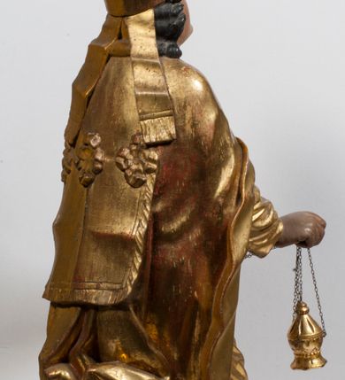 Zdjęcie nr 11: Tabernakulum w formie Arki Przymierza, adorowanej przez dwie klęczące figury starotestamentowego arcykapłana i biskupa oraz dwie stojące postacie lewitów oraz klęczące aniołów, zwieńczonej glorią promienistą z okiem Opatrzności. Wsparte na ośmiu cylindrycznych nóżkach prostopadłościenne tabernakulum jest od frontu podzielone na trzy pola wyznaczone kanelowanymi listwami. Pole środkowe stanowią drzwiczki z rytymi cyframi Dekalogu z ukrzyżowanym Chrystusem, pola boczne z wiązką pałek wodnych z jednej i winnym gronem z drugiej strony. Na skłębionych chmurach z podestem leży Baranek (obecnie w jego miejscu znajduje się figura Matki Boskiej). W tle znacznej wielkości gloria promienista z wieńcem chmur i umieszczonym w nim okiem Opatrzności oraz dwie uskrzydlone główki, nad którymi papieska tiara ze skrzyżowanymi kluczami. Po bokach Baranka dwa klęczące, uskrzydlone nagie aniołki. Obok tabernakulum dwie klęczące postacie z kadzielnicami w rękach. Postać po lewej w stroju biskupim, w albie, stule i kapie oraz infule z dwiema taśmami. Postać po prawej stronie jest ubrany w tradycyjny starotestamentowy strój – efod czyli tunikę i płaszcz wierzchni przepasany na biodrach, pektorał na piersi i zawój na głowie. Za nimi dwaj stojący lewici w tunikach przepasanych na biodrach i wysokich butach. Adorujące Baranka anioły w pozycji klęczącej z rękami skrzyżowanymi na piersi, ze skrzydłami na plecach i nogami w połowie utworzonymi ze skrzydeł.  


