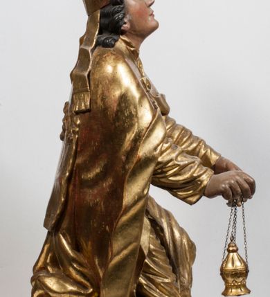 Zdjęcie nr 14: Tabernakulum w formie Arki Przymierza, adorowanej przez dwie klęczące figury starotestamentowego arcykapłana i biskupa oraz dwie stojące postacie lewitów oraz klęczące aniołów, zwieńczonej glorią promienistą z okiem Opatrzności. Wsparte na ośmiu cylindrycznych nóżkach prostopadłościenne tabernakulum jest od frontu podzielone na trzy pola wyznaczone kanelowanymi listwami. Pole środkowe stanowią drzwiczki z rytymi cyframi Dekalogu z ukrzyżowanym Chrystusem, pola boczne z wiązką pałek wodnych z jednej i winnym gronem z drugiej strony. Na skłębionych chmurach z podestem leży Baranek (obecnie w jego miejscu znajduje się figura Matki Boskiej). W tle znacznej wielkości gloria promienista z wieńcem chmur i umieszczonym w nim okiem Opatrzności oraz dwie uskrzydlone główki, nad którymi papieska tiara ze skrzyżowanymi kluczami. Po bokach Baranka dwa klęczące, uskrzydlone nagie aniołki. Obok tabernakulum dwie klęczące postacie z kadzielnicami w rękach. Postać po lewej w stroju biskupim, w albie, stule i kapie oraz infule z dwiema taśmami. Postać po prawej stronie jest ubrany w tradycyjny starotestamentowy strój – efod czyli tunikę i płaszcz wierzchni przepasany na biodrach, pektorał na piersi i zawój na głowie. Za nimi dwaj stojący lewici w tunikach przepasanych na biodrach i wysokich butach. Adorujące Baranka anioły w pozycji klęczącej z rękami skrzyżowanymi na piersi, ze skrzydłami na plecach i nogami w połowie utworzonymi ze skrzydeł.  


