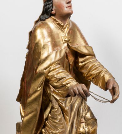 Zdjęcie nr 16: Tabernakulum w formie Arki Przymierza, adorowanej przez dwie klęczące figury starotestamentowego arcykapłana i biskupa oraz dwie stojące postacie lewitów oraz klęczące aniołów, zwieńczonej glorią promienistą z okiem Opatrzności. Wsparte na ośmiu cylindrycznych nóżkach prostopadłościenne tabernakulum jest od frontu podzielone na trzy pola wyznaczone kanelowanymi listwami. Pole środkowe stanowią drzwiczki z rytymi cyframi Dekalogu z ukrzyżowanym Chrystusem, pola boczne z wiązką pałek wodnych z jednej i winnym gronem z drugiej strony. Na skłębionych chmurach z podestem leży Baranek (obecnie w jego miejscu znajduje się figura Matki Boskiej). W tle znacznej wielkości gloria promienista z wieńcem chmur i umieszczonym w nim okiem Opatrzności oraz dwie uskrzydlone główki, nad którymi papieska tiara ze skrzyżowanymi kluczami. Po bokach Baranka dwa klęczące, uskrzydlone nagie aniołki. Obok tabernakulum dwie klęczące postacie z kadzielnicami w rękach. Postać po lewej w stroju biskupim, w albie, stule i kapie oraz infule z dwiema taśmami. Postać po prawej stronie jest ubrany w tradycyjny starotestamentowy strój – efod czyli tunikę i płaszcz wierzchni przepasany na biodrach, pektorał na piersi i zawój na głowie. Za nimi dwaj stojący lewici w tunikach przepasanych na biodrach i wysokich butach. Adorujące Baranka anioły w pozycji klęczącej z rękami skrzyżowanymi na piersi, ze skrzydłami na plecach i nogami w połowie utworzonymi ze skrzydeł.  


