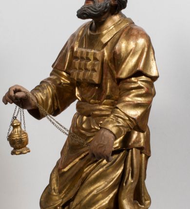 Zdjęcie nr 21: Tabernakulum w formie Arki Przymierza, adorowanej przez dwie klęczące figury starotestamentowego arcykapłana i biskupa oraz dwie stojące postacie lewitów oraz klęczące aniołów, zwieńczonej glorią promienistą z okiem Opatrzności. Wsparte na ośmiu cylindrycznych nóżkach prostopadłościenne tabernakulum jest od frontu podzielone na trzy pola wyznaczone kanelowanymi listwami. Pole środkowe stanowią drzwiczki z rytymi cyframi Dekalogu z ukrzyżowanym Chrystusem, pola boczne z wiązką pałek wodnych z jednej i winnym gronem z drugiej strony. Na skłębionych chmurach z podestem leży Baranek (obecnie w jego miejscu znajduje się figura Matki Boskiej). W tle znacznej wielkości gloria promienista z wieńcem chmur i umieszczonym w nim okiem Opatrzności oraz dwie uskrzydlone główki, nad którymi papieska tiara ze skrzyżowanymi kluczami. Po bokach Baranka dwa klęczące, uskrzydlone nagie aniołki. Obok tabernakulum dwie klęczące postacie z kadzielnicami w rękach. Postać po lewej w stroju biskupim, w albie, stule i kapie oraz infule z dwiema taśmami. Postać po prawej stronie jest ubrany w tradycyjny starotestamentowy strój – efod czyli tunikę i płaszcz wierzchni przepasany na biodrach, pektorał na piersi i zawój na głowie. Za nimi dwaj stojący lewici w tunikach przepasanych na biodrach i wysokich butach. Adorujące Baranka anioły w pozycji klęczącej z rękami skrzyżowanymi na piersi, ze skrzydłami na plecach i nogami w połowie utworzonymi ze skrzydeł.  


